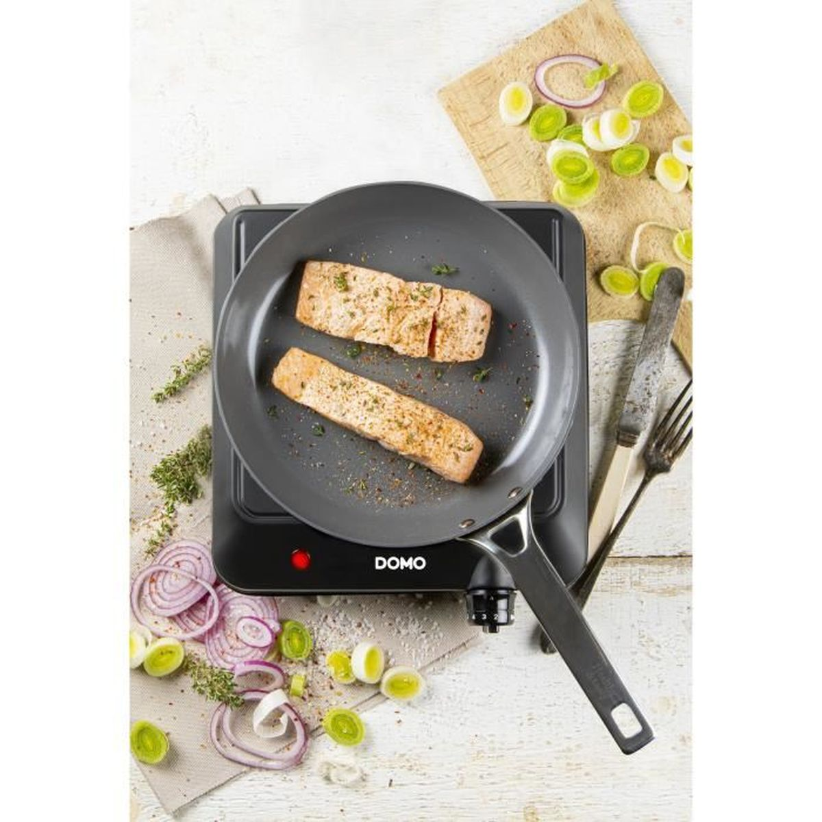 Pfanne mit gekochtem Lachs auf einem schwarzen Herd. Umgeben von geschnittenen Zwiebeln und Kräutern. Weiße Oberfläche.
