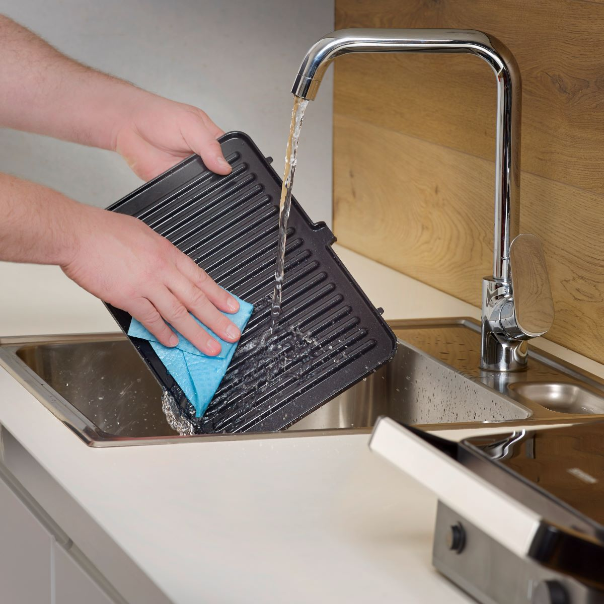 A person washing a black grill pan under running water in a kitchen sink.