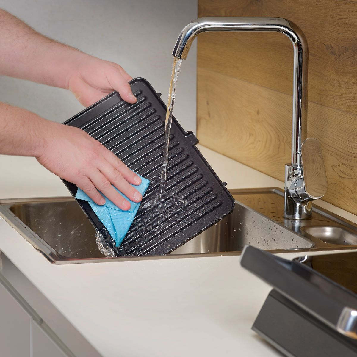Person wäscht eine Grillpfanne in einem Waschbecken mit blauem Tuch und fließendem Wasser.