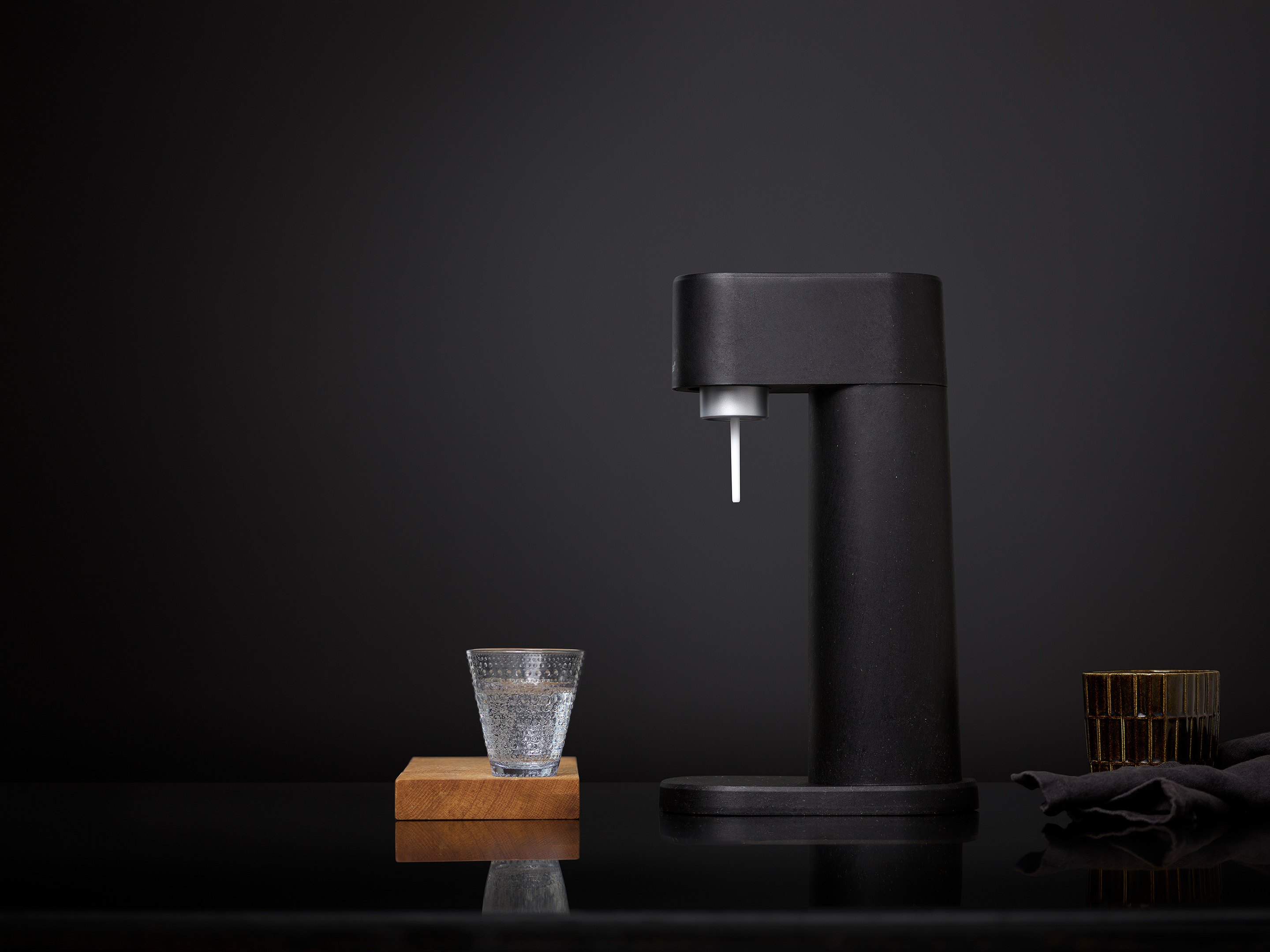Sifone nero con bicchiere d'acqua su supporto in legno e sfondo scuro.