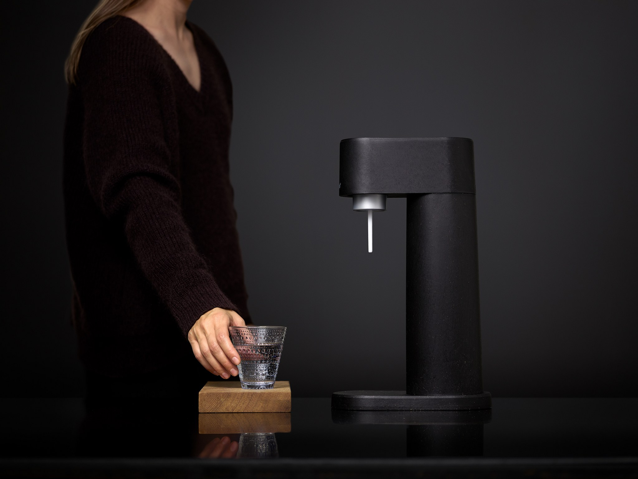 Una persona versa acqua da un produttore di soda nero in un bicchiere d'acqua su un supporto di legno.