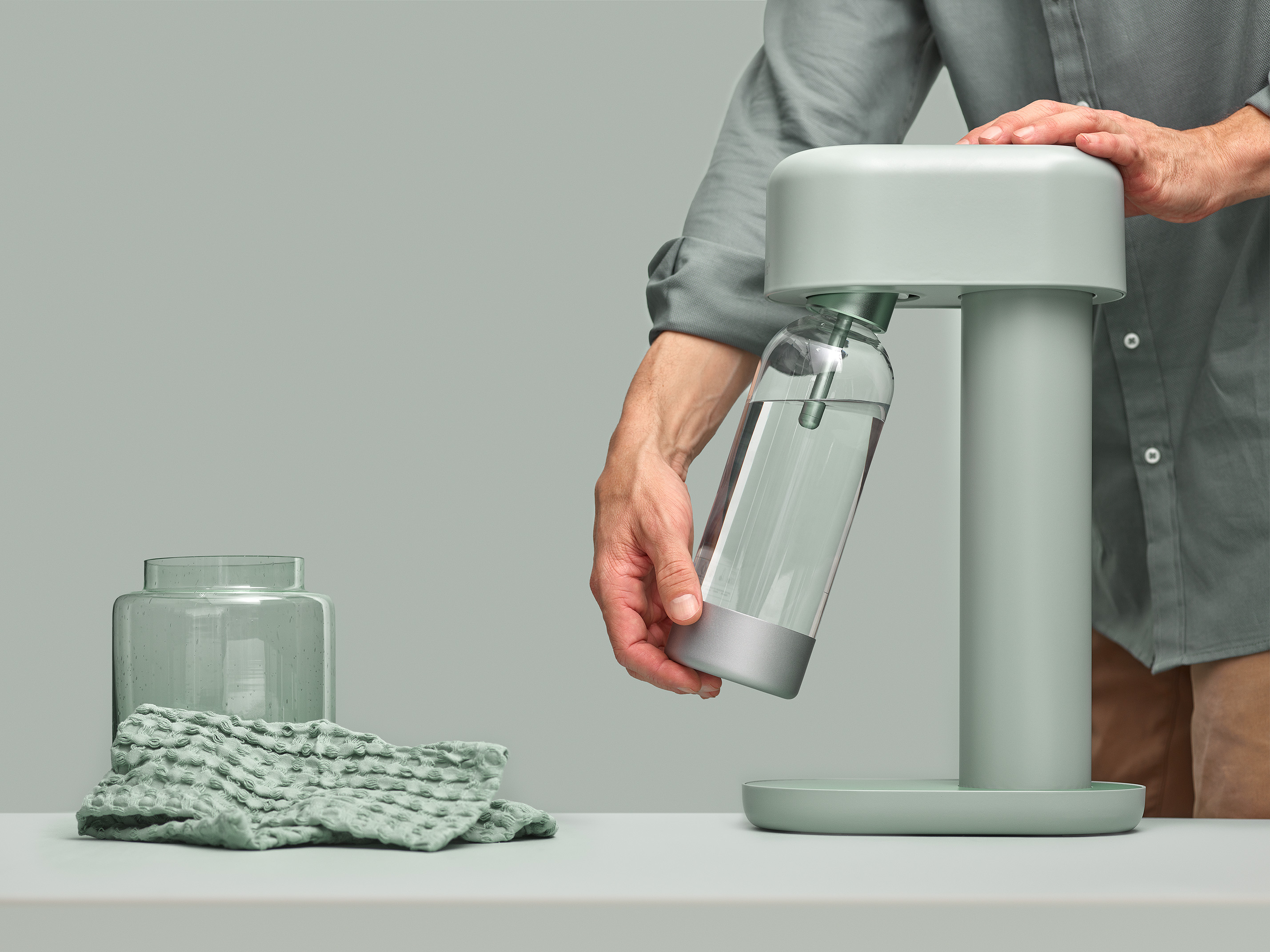 A light green soda maker with a glass bottle. A person is holding and inserting the bottle.