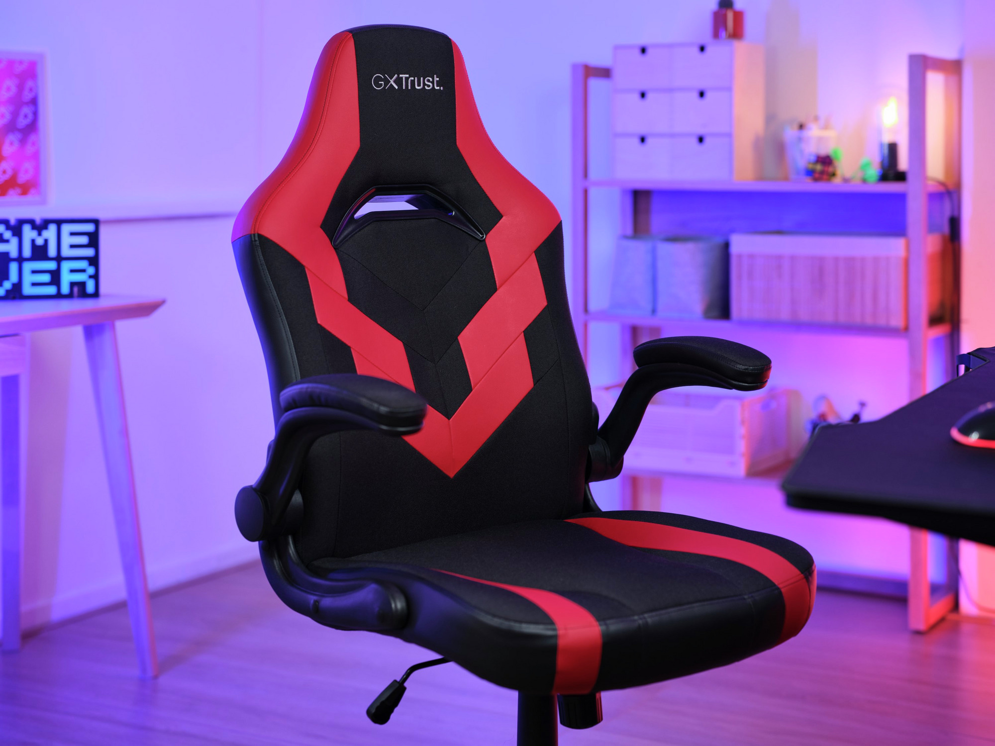 Een zwarte en rode gamingstoel met 'GXtrust' logo. Achtergrond is een bureau en planken met diverse items.