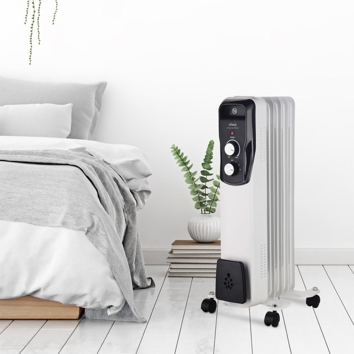 Radiador de aceite blanco en una habitación con cama, planta y suelo de madera.