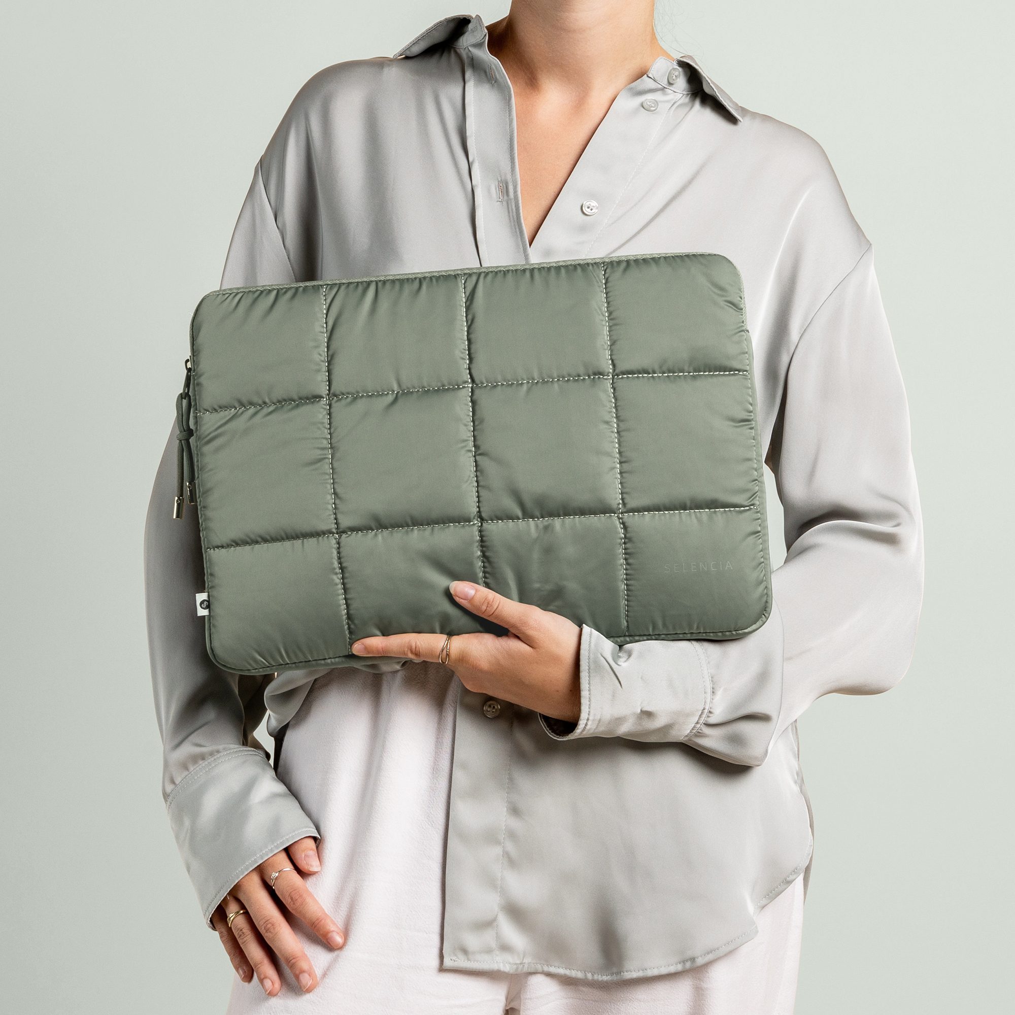 Woman holding a green quilted laptop sleeve. She wears a gray shirt and white pants.