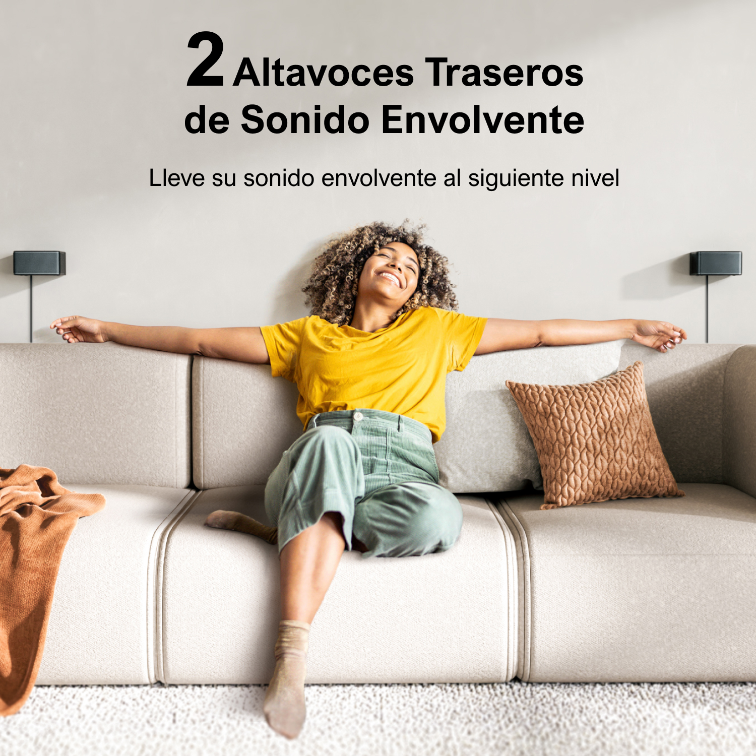 Mujer en sofá beige con brazos extendidos, dos altavoces en la pared. Sonriendo, camisa amarilla, pantalón verde.