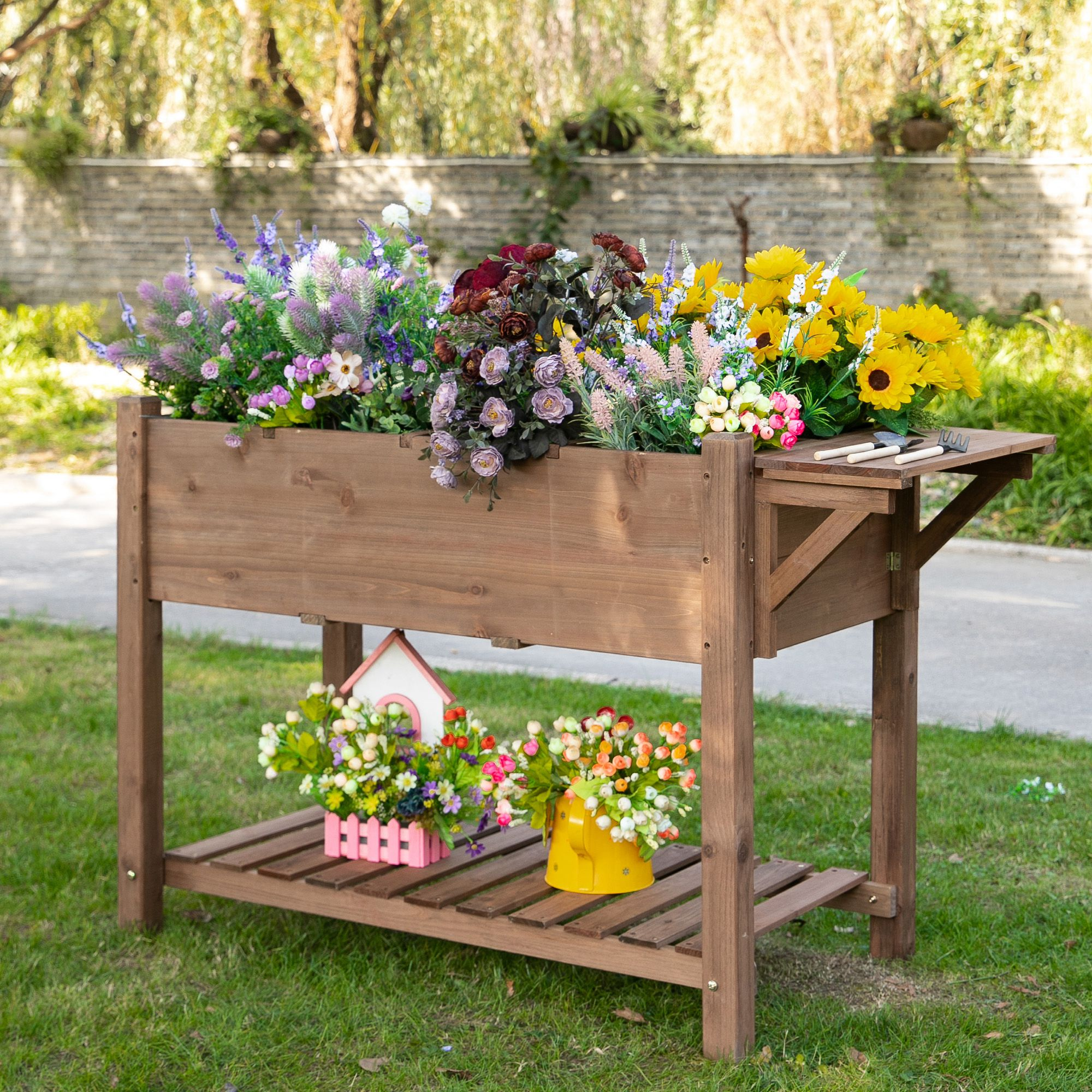 Aiuola rialzata in legno con fiori colorati, una casetta e attrezzi su un prato verde.