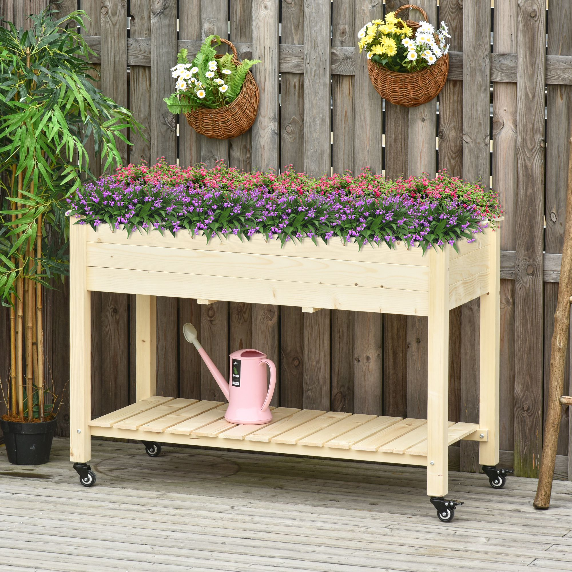 Fioriera in legno su ruote con fiori e cesti. Annaffiatoio rosa. Deck in legno e recinzione sullo sfondo.