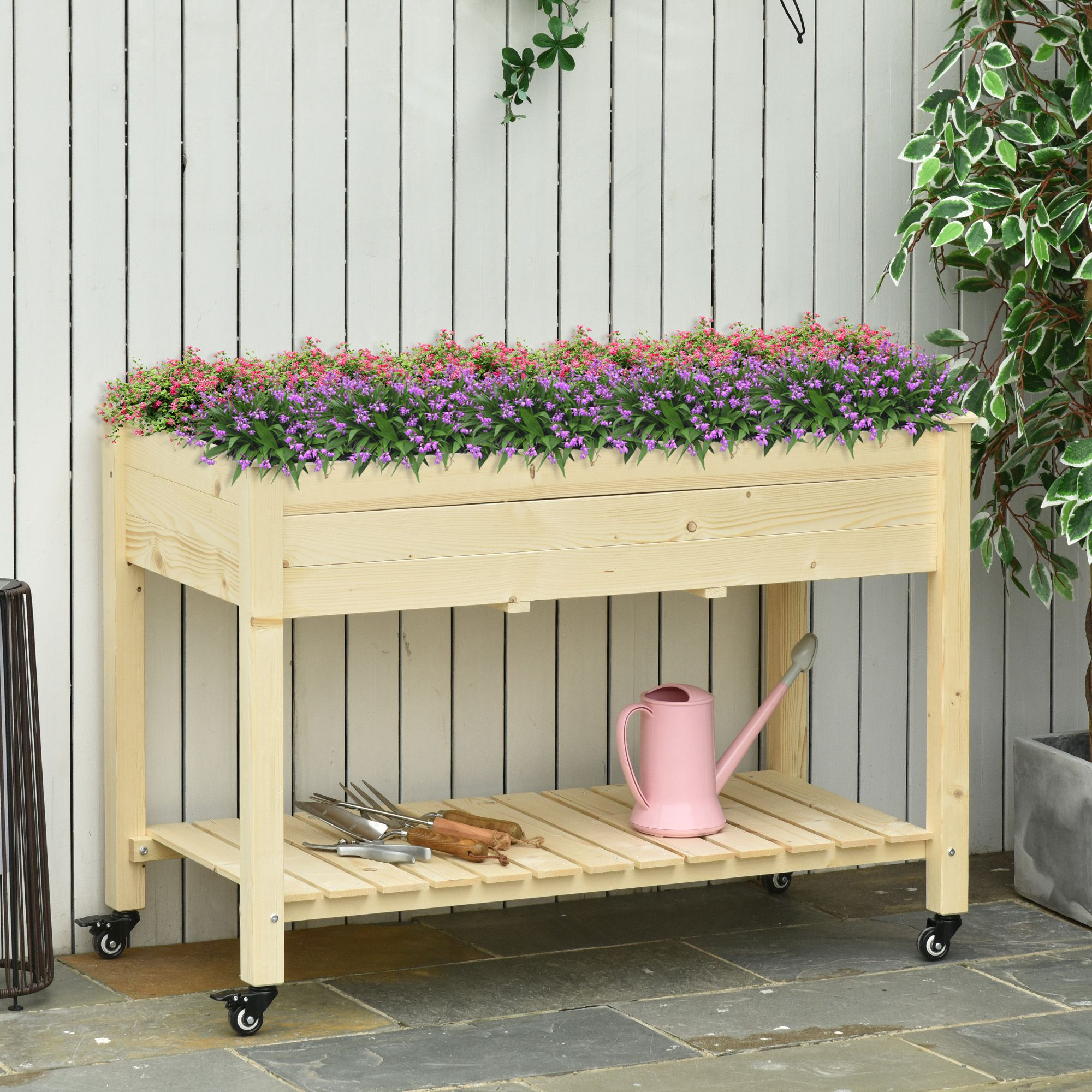 Fioriera in legno con fiori, su ruote, vicino a una staccionata bianca.