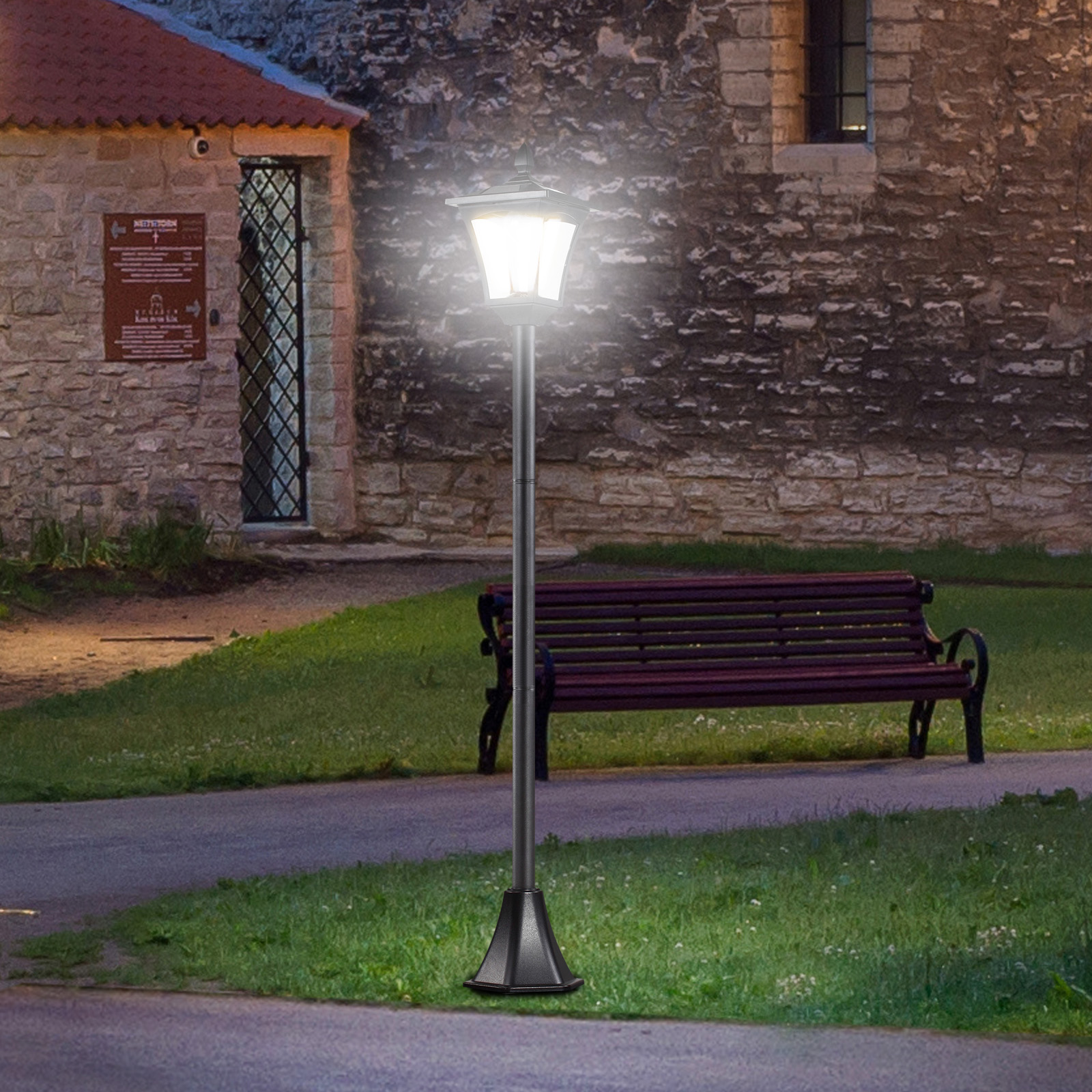 Un lampione illuminato in un parco, con una panchina e un edificio in pietra.