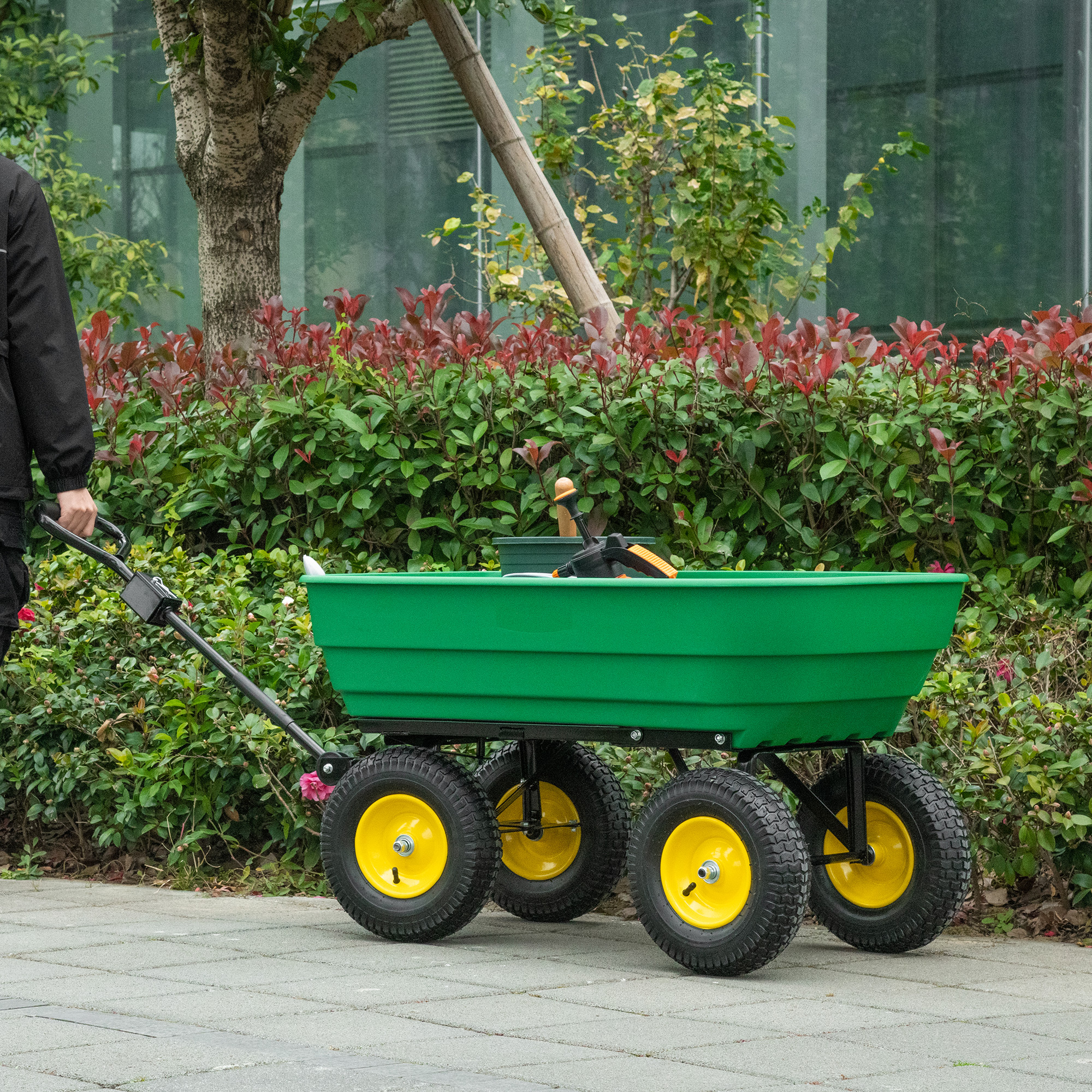 Una persona che tira un carrello da giardino verde con ruote gialle su una superficie pavimentata con fogliame.