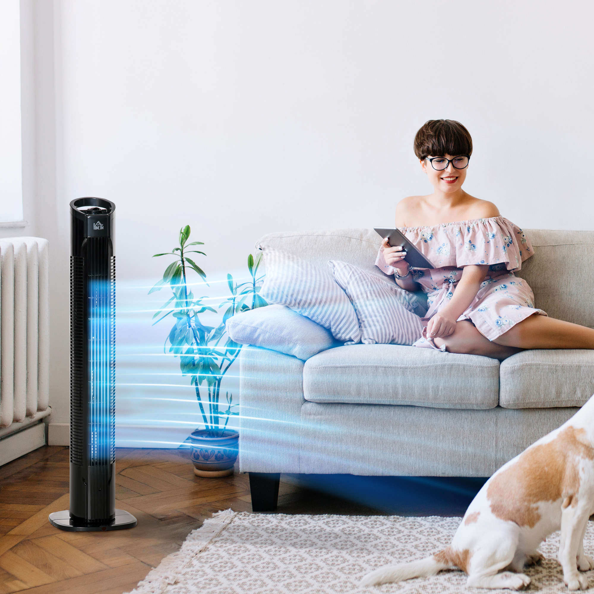 Una donna su un divano con un tablet, un ventilatore a torre nero a sinistra. Un cane a destra.