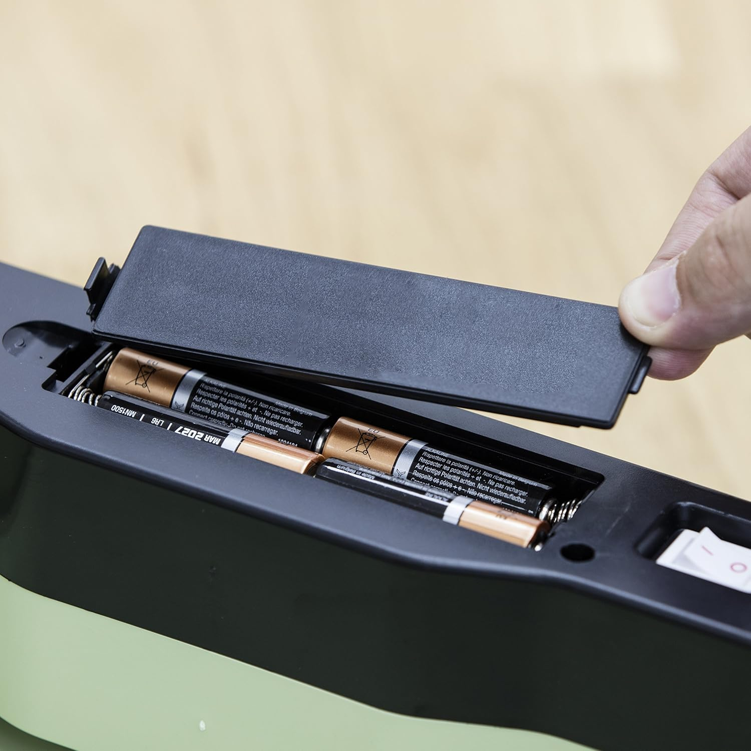 Hand öffnet ein Fach und zeigt 4 AA-Batterien in einem schwarz-grünen Gerät.