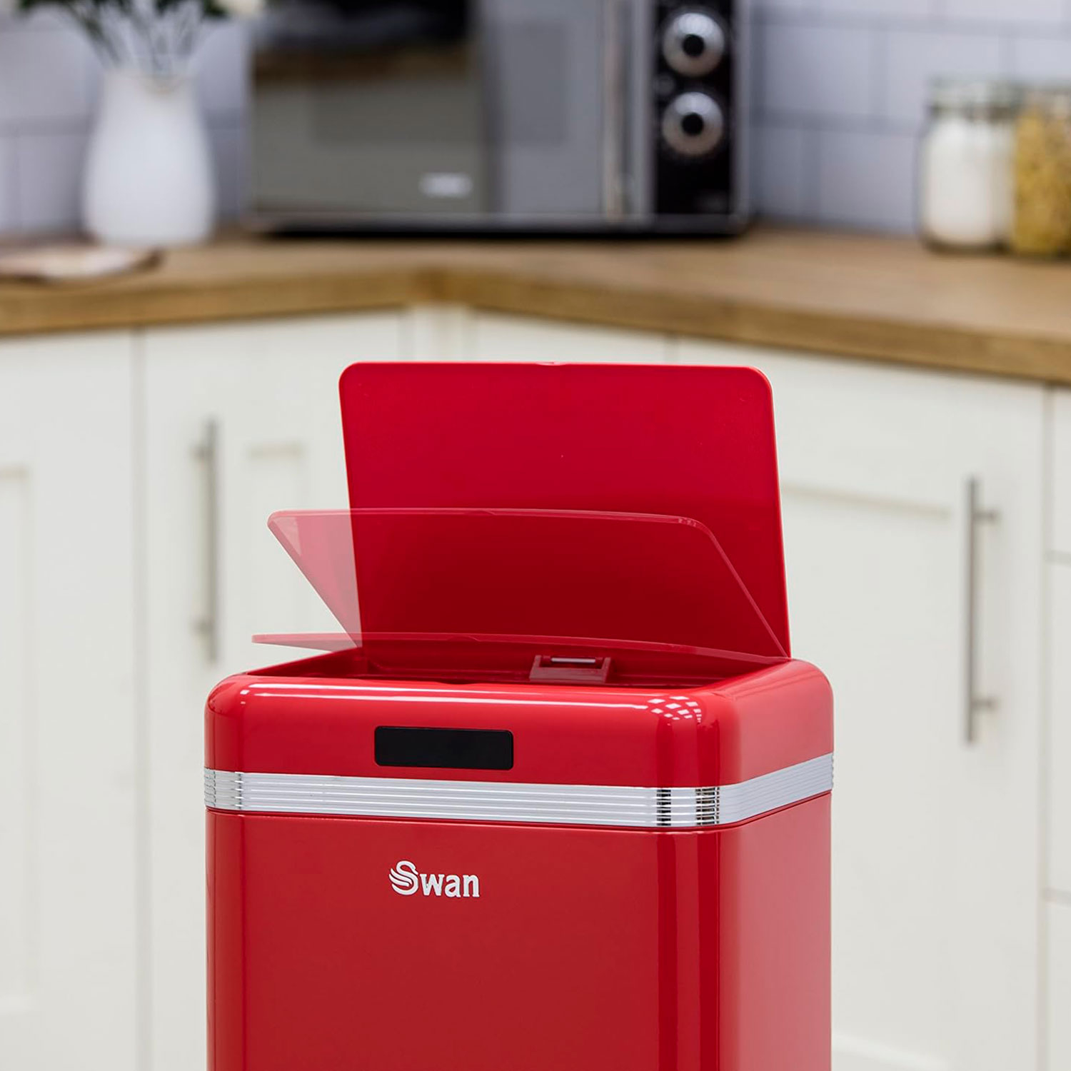 Contenedor de basura rojo con tapa abierta en una cocina, gabinetes blancos y microondas al fondo.