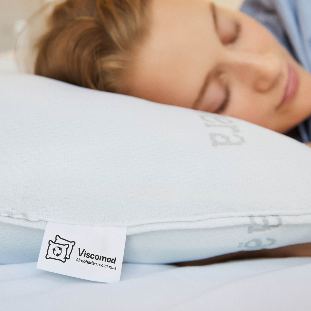 Mujer durmiendo en una almohada azul claro con una etiqueta Viscomed, que dice 'Almohadas recicladas'.