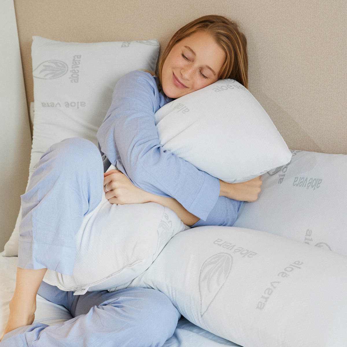Una mujer con pijama azul abraza almohadas blancas en una cama. Las almohadas tienen la palabra 'aloe vera'.