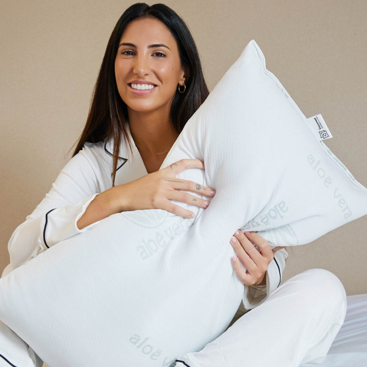 Mujer en pijama sosteniendo una almohada blanca. Sonríe a la cámara en una cama.