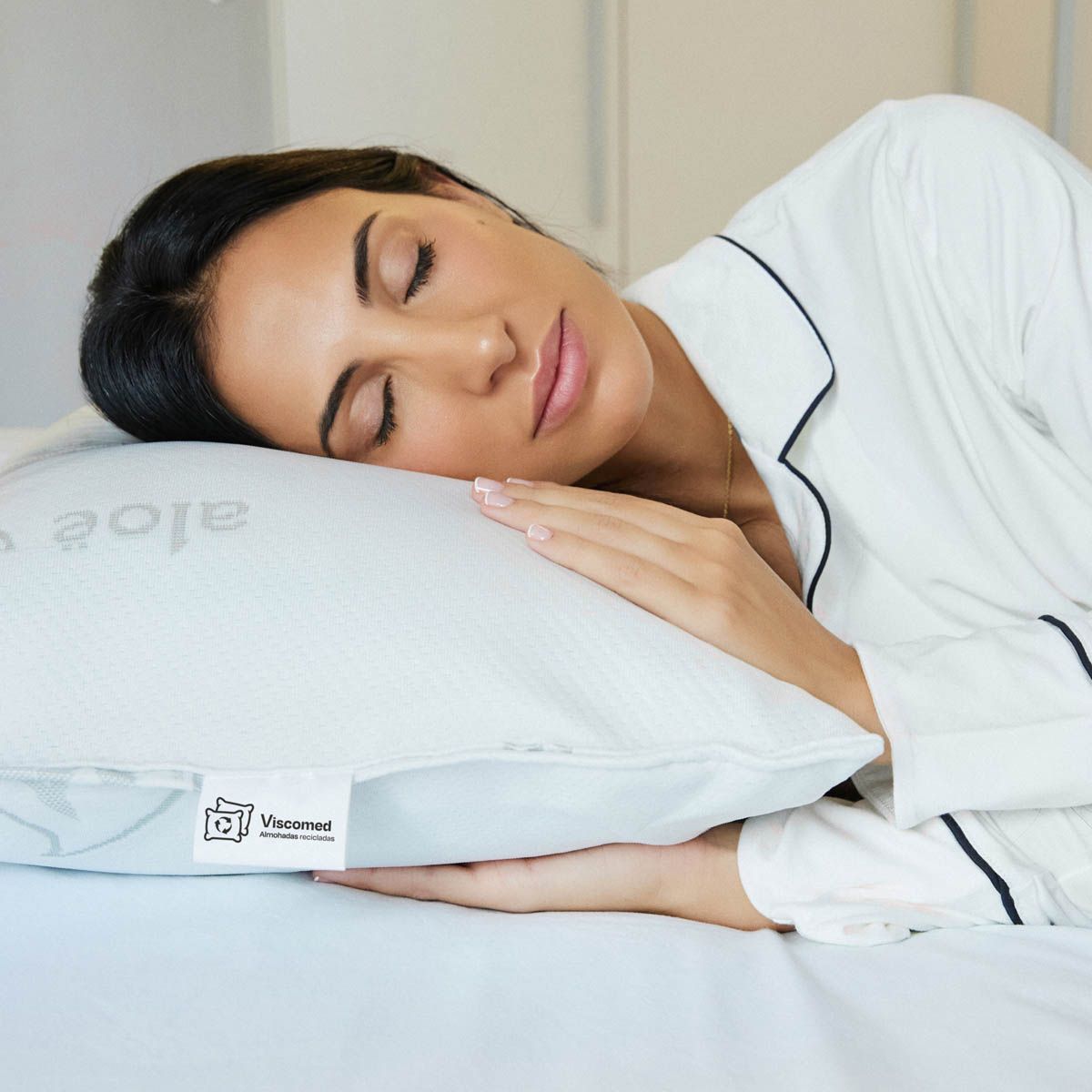 Mujer durmiendo en una almohada blanca. La almohada tiene una etiqueta Viscomed. Viste un pijama blanco.