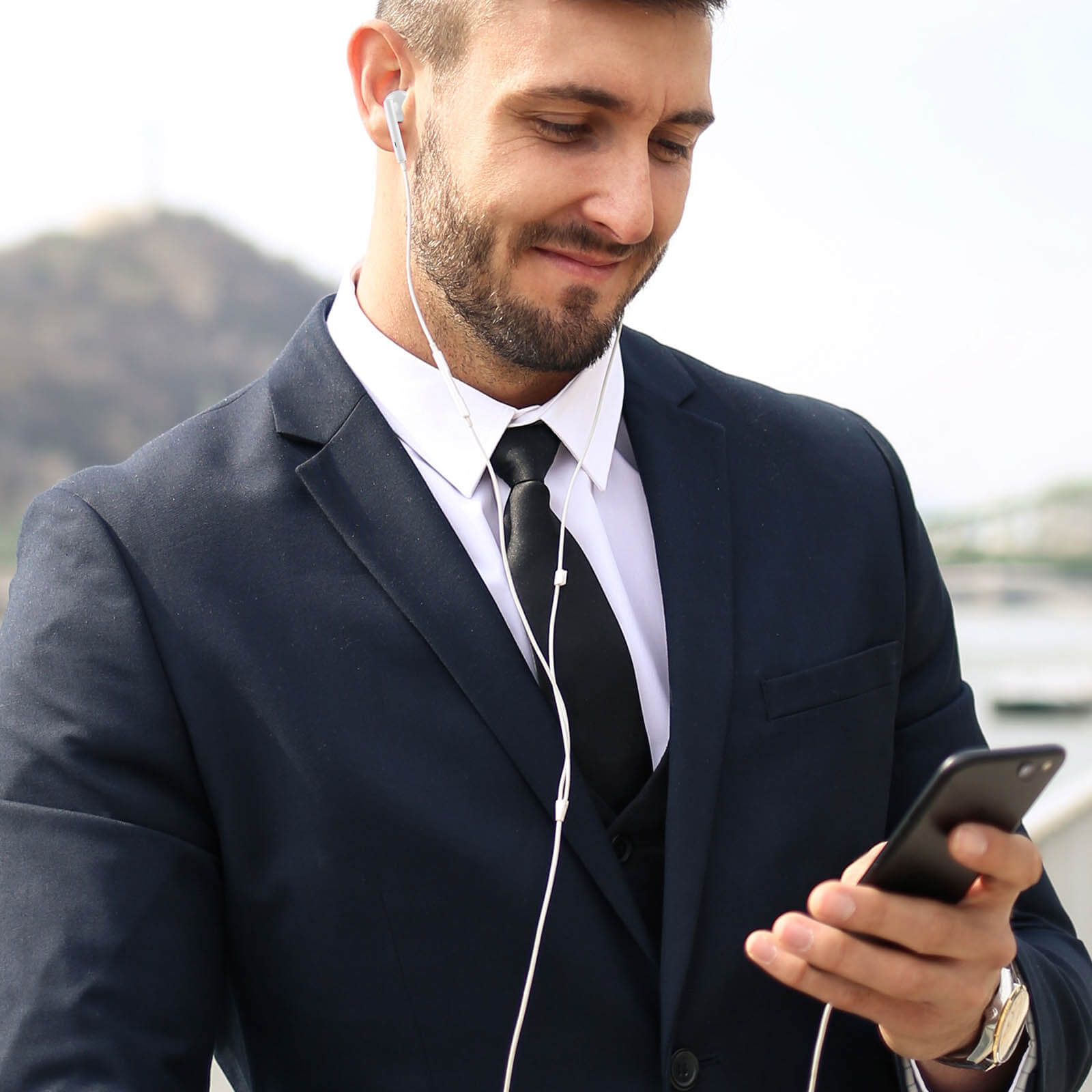 Un hombre en traje sonríe, escuchando música con auriculares blancos mientras mira su teléfono.