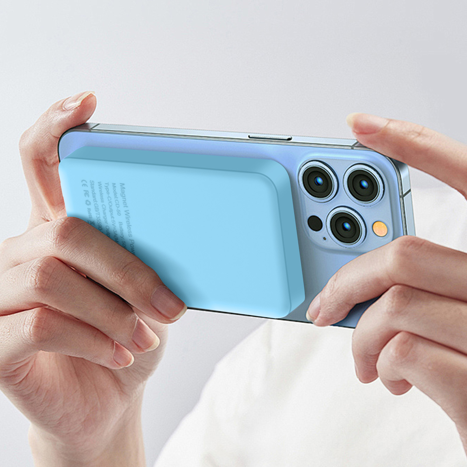 Handen die een blauwe telefoon vasthouden met een lichtblauwe magnetische draadloze powerbank eraan.
