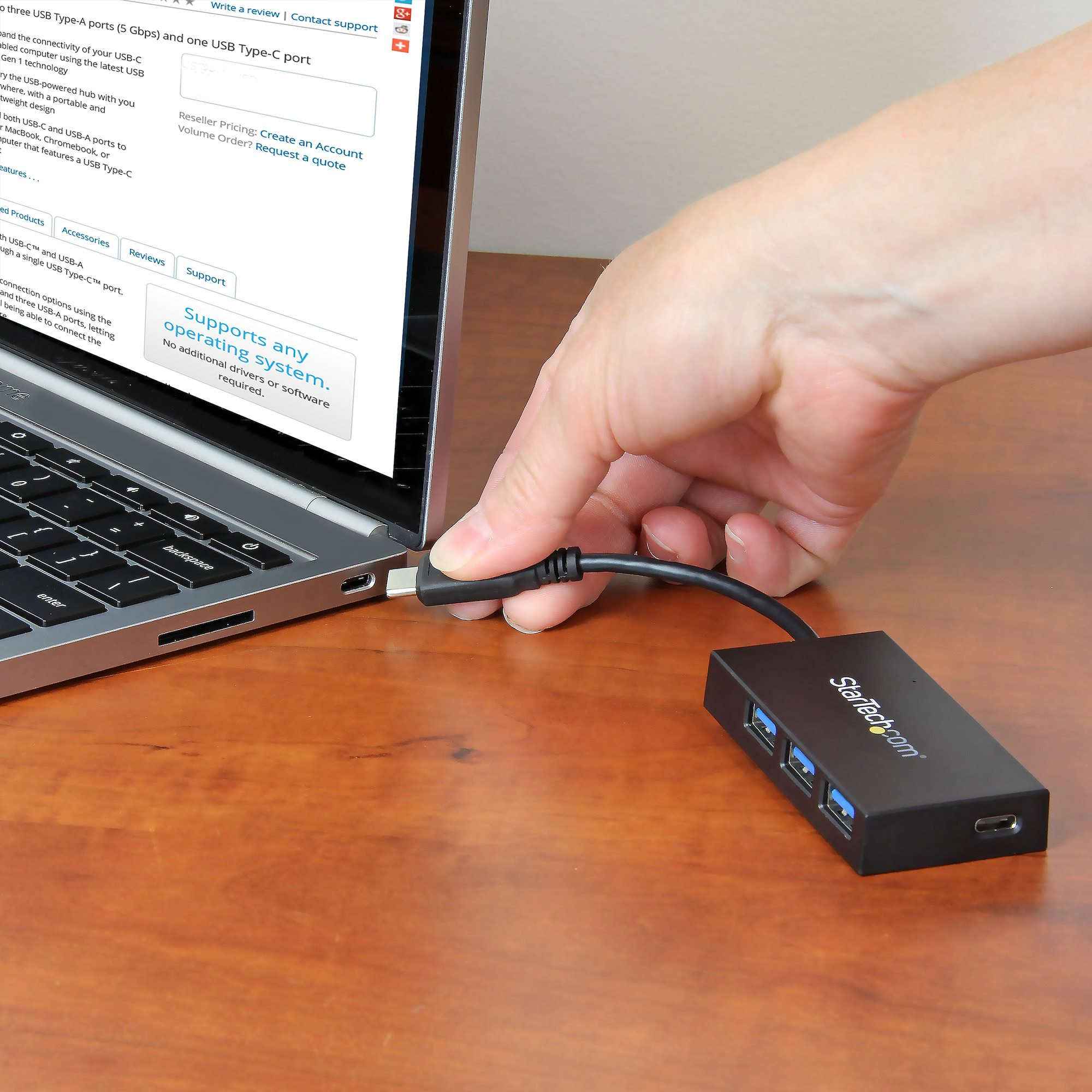Una mano che collega un cavo USB-C a un laptop, con un hub USB collegato, su una scrivania di legno.