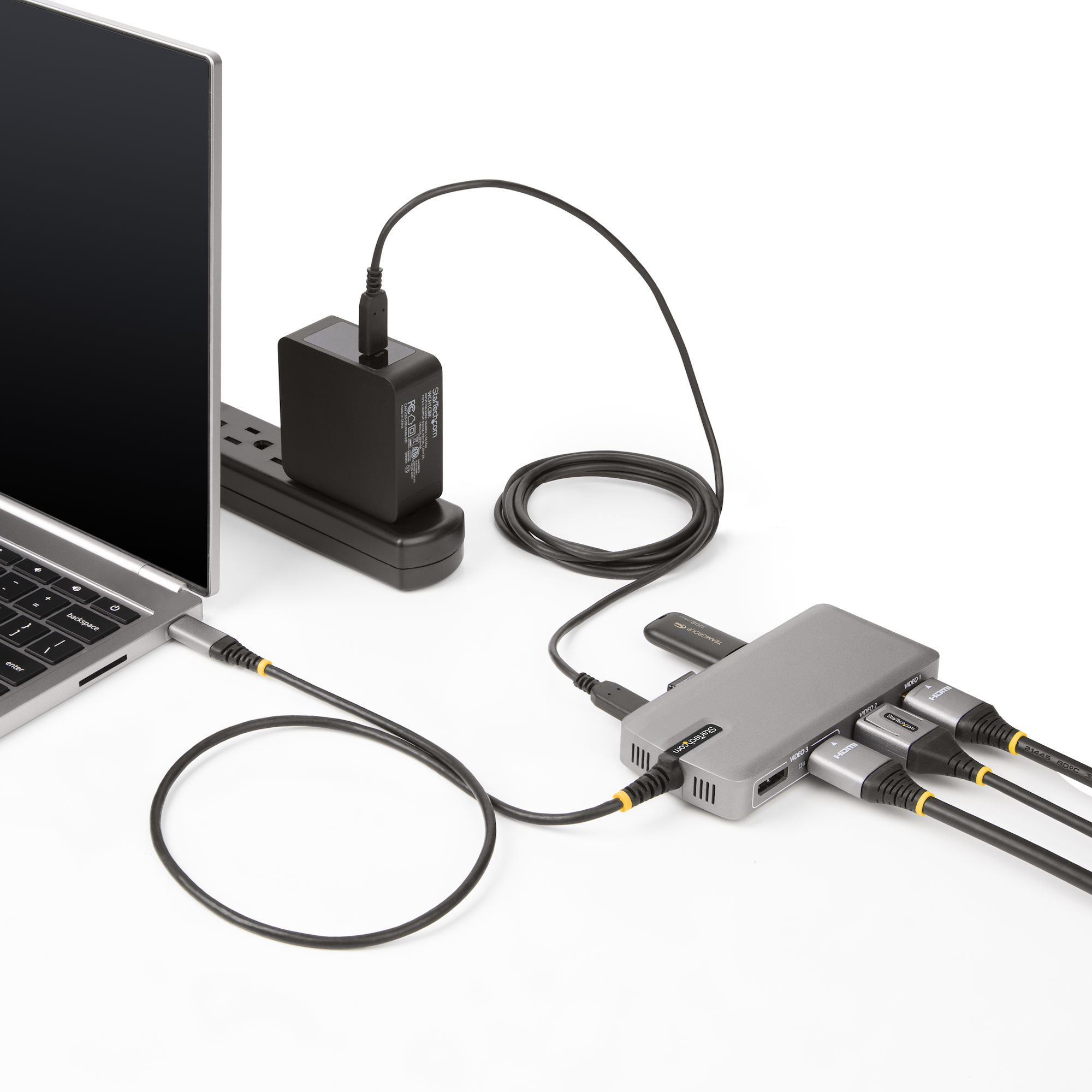 Laptop verbunden mit Netzteil und grauem Hub mit mehreren Anschlüssen, auf weißer Oberfläche.