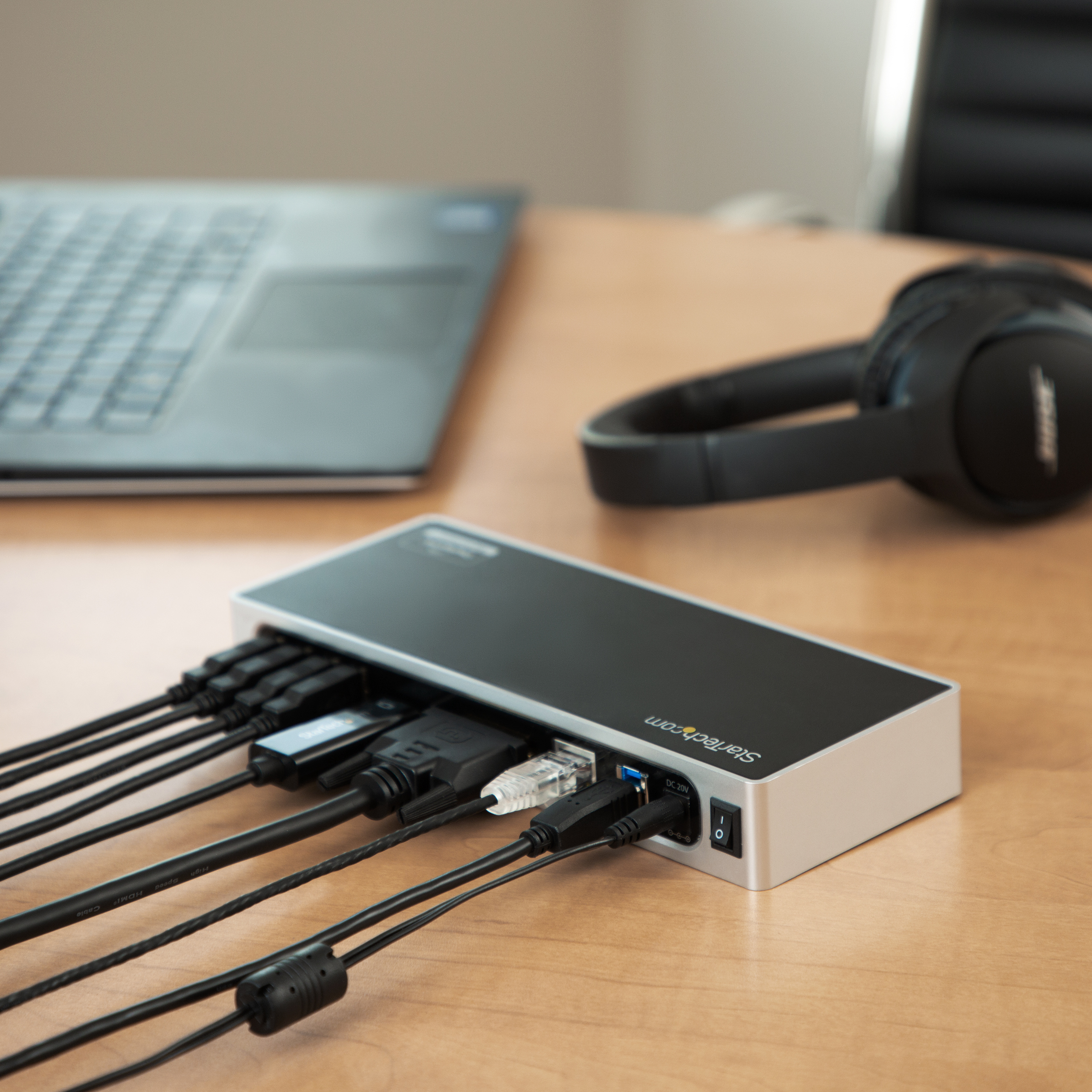Docking station con cavi collegati, laptop e cuffie su una scrivania in legno.