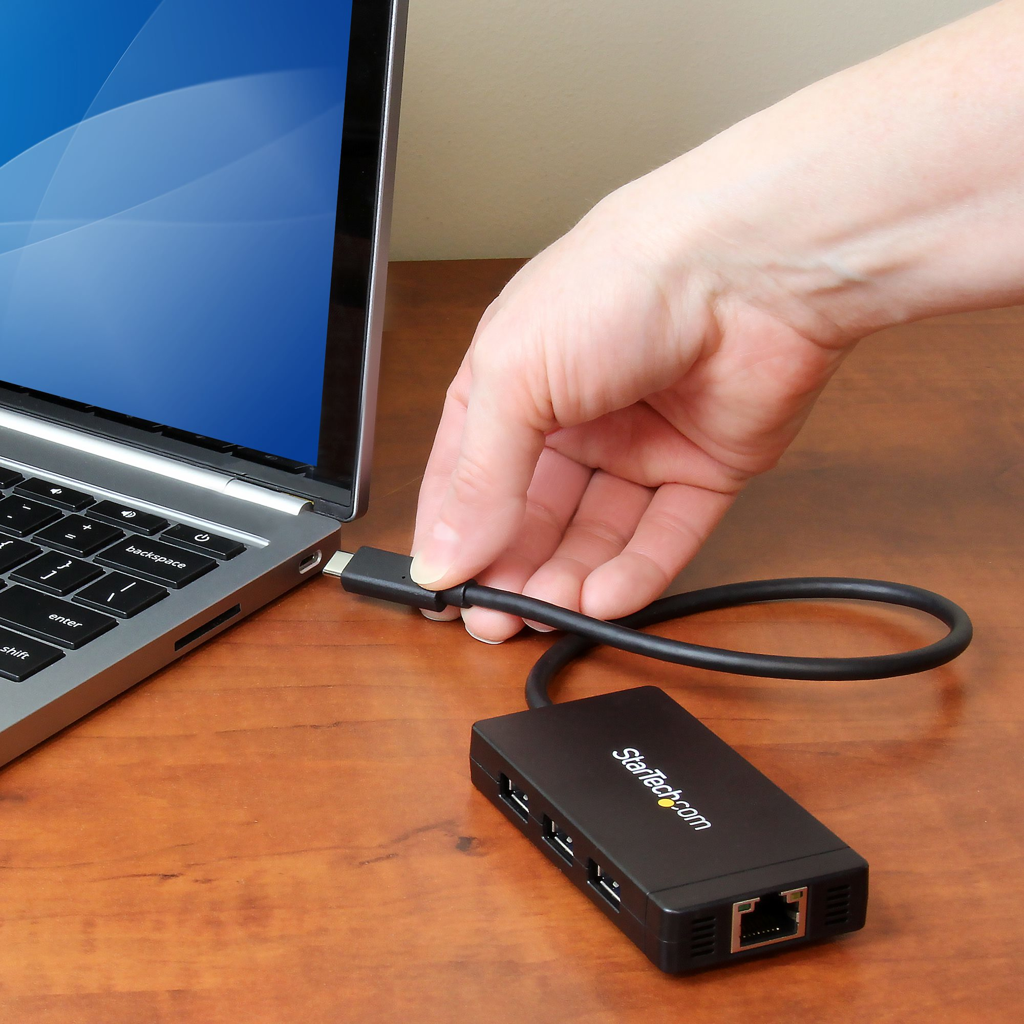Una mano conecta un concentrador USB-C negro a una laptop. El concentrador está en un escritorio de madera.