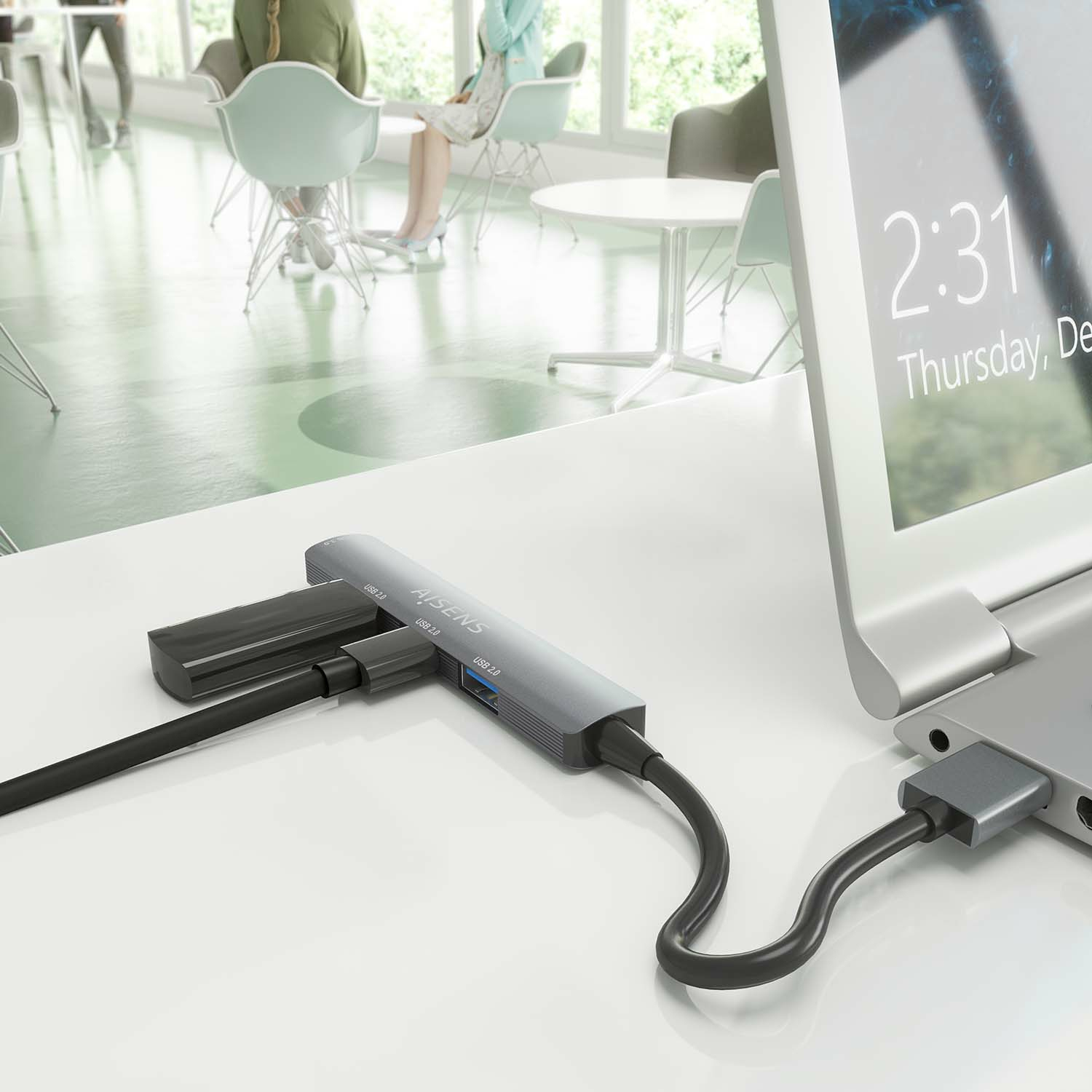 Laptop collegato a un hub USB grigio su una scrivania bianca. Persone sfocate.