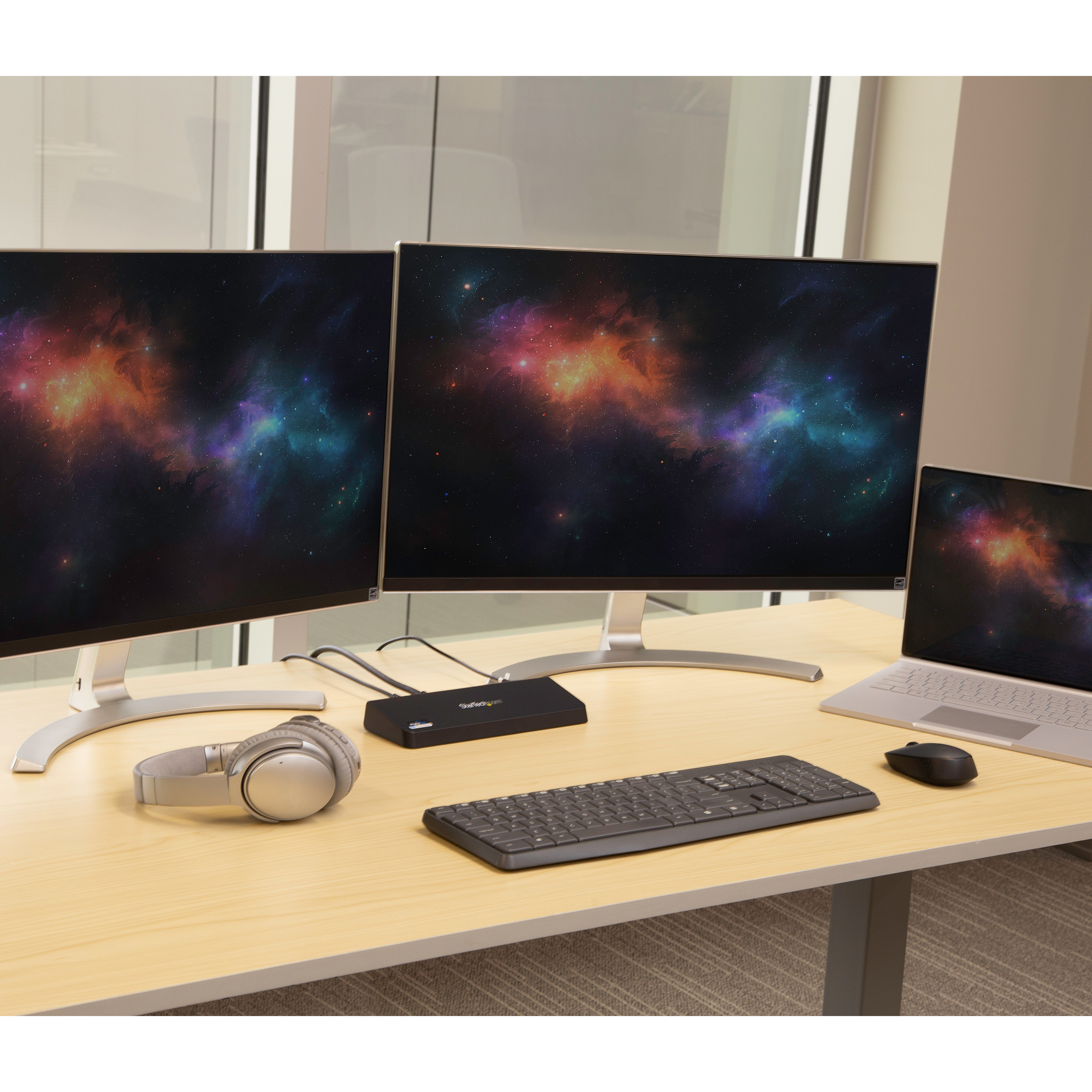 Escritorio de oficina con dos monitores, portátil, teclado, auriculares y concentrador. Fondo: ventana.