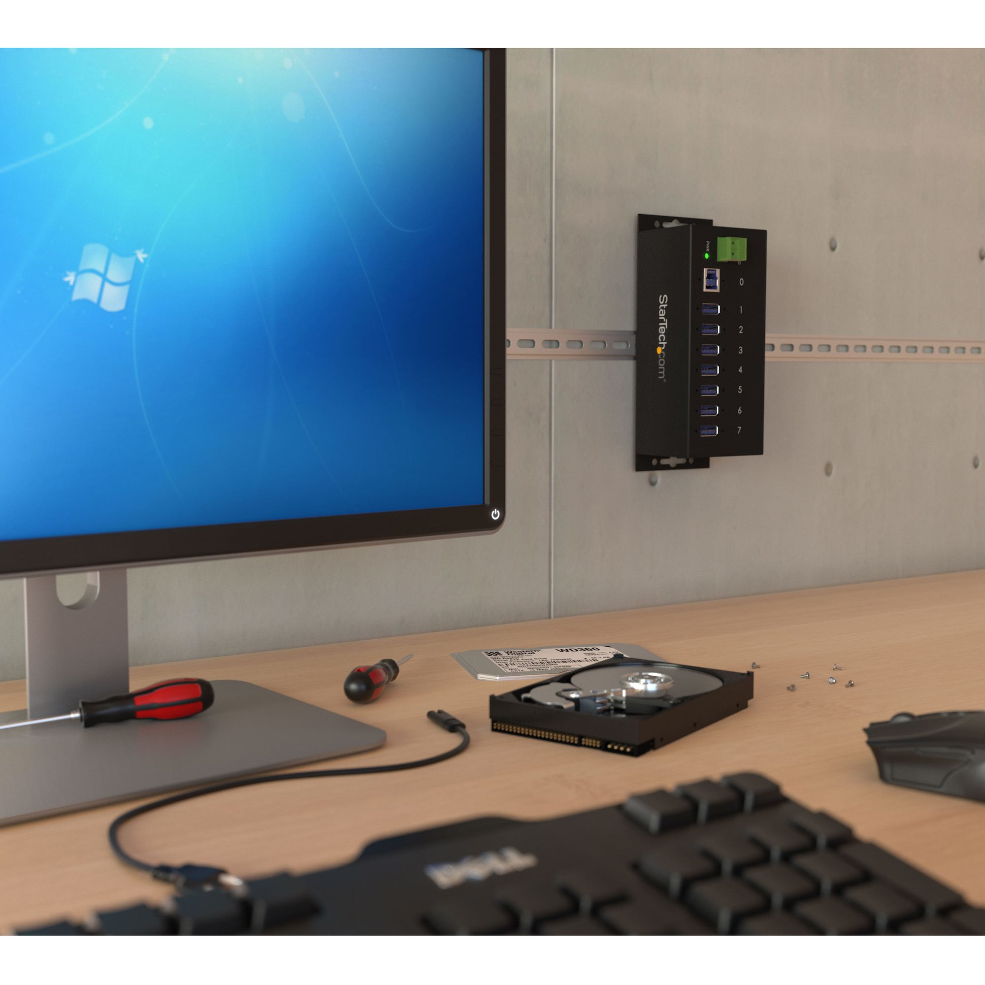 Een computermonitor, een toetsenbord en een zwarte USB-hub aan de muur, met gereedschap op het bureau.