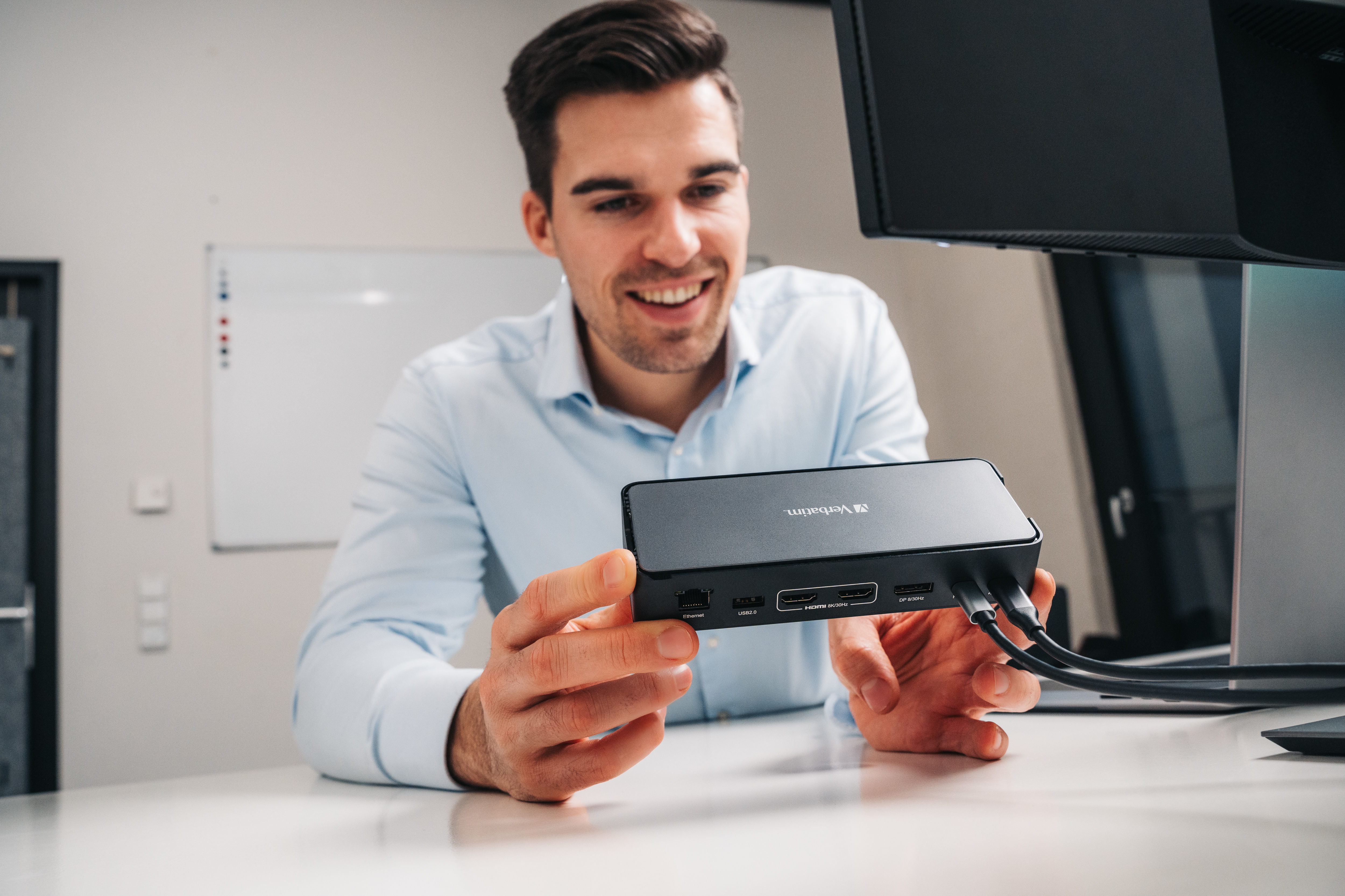 Mann hält ein schwarzes Verbatim-Gerät. Weißer Schreibtisch, Monitor und Whiteboard im Hintergrund.