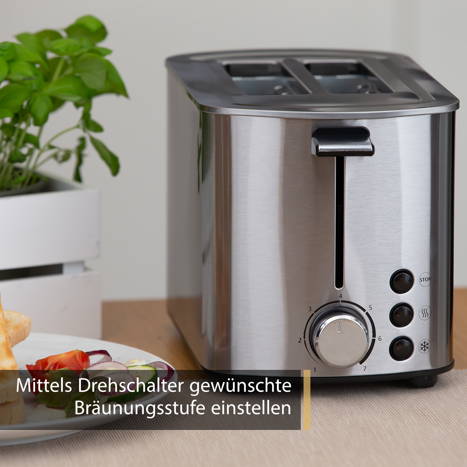 Toaster aus Edelstahl auf einem Holztisch. Teller mit Essen und Pflanze im Hintergrund. Text ist Deutsch.