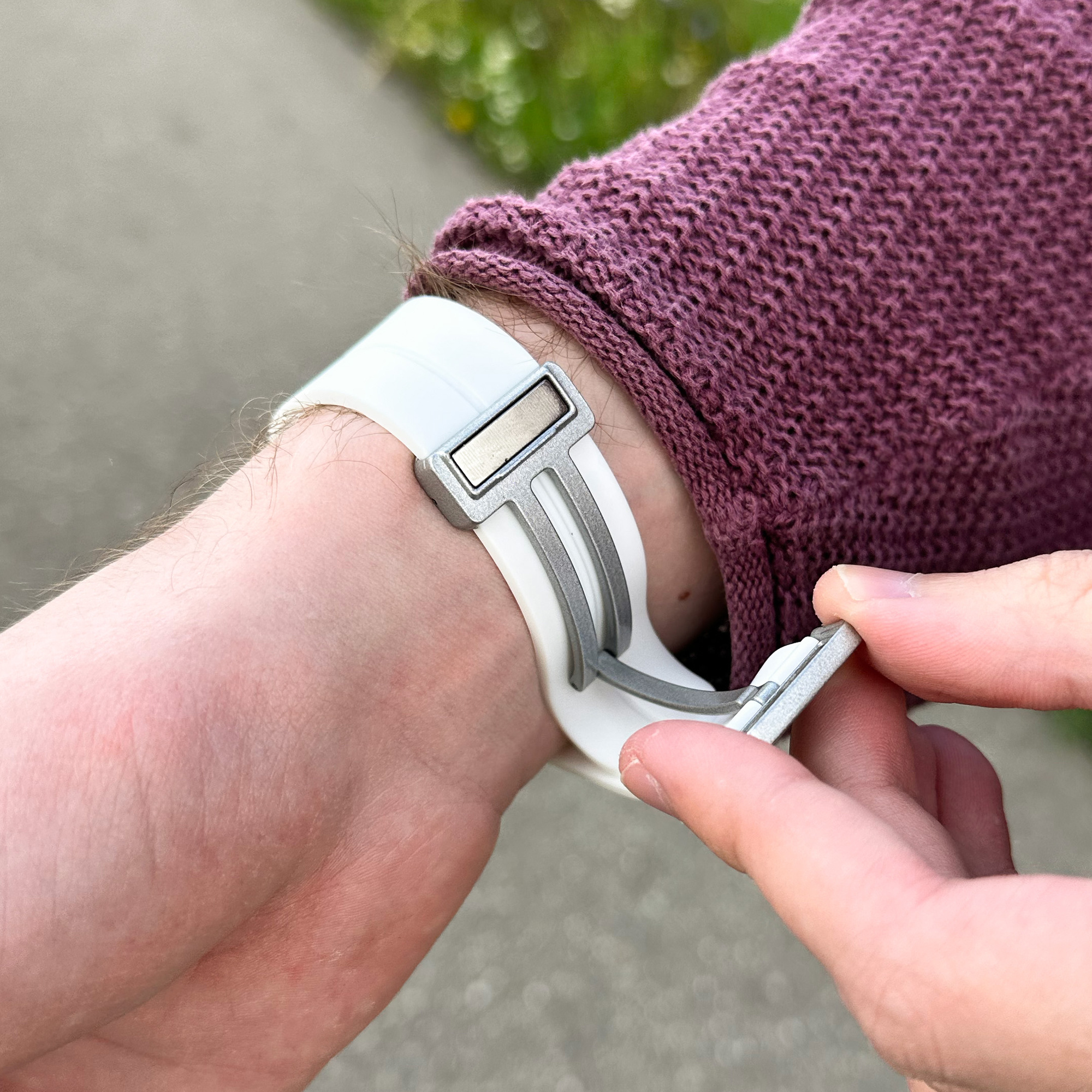 Das Handgelenk einer Person mit einem weißen Uhrenarmband, der Verschluss wird geöffnet, verschwommener Hintergrund.