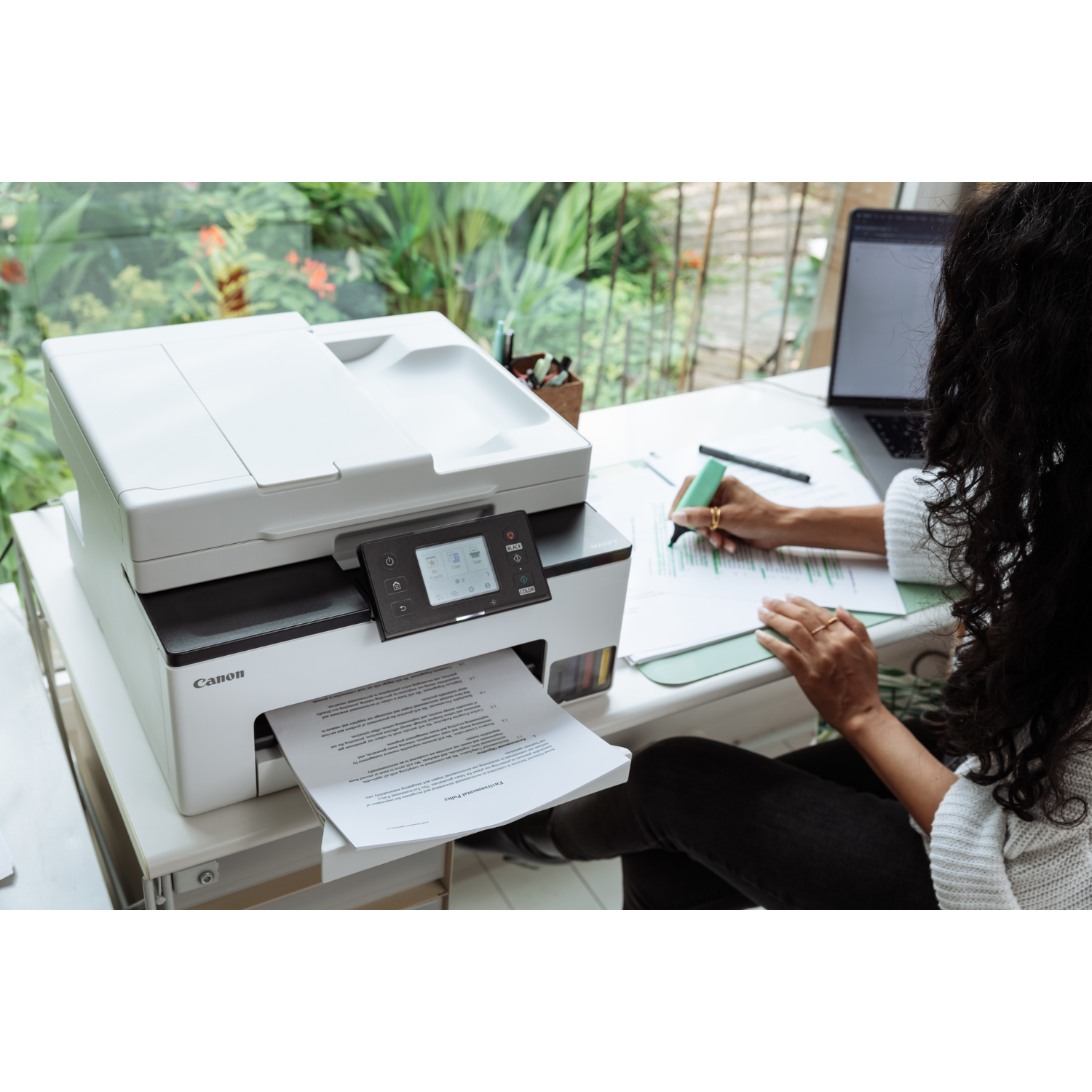 Kobieta używająca drukarki Canon przy biurku, z laptopem i oknem w tle.
