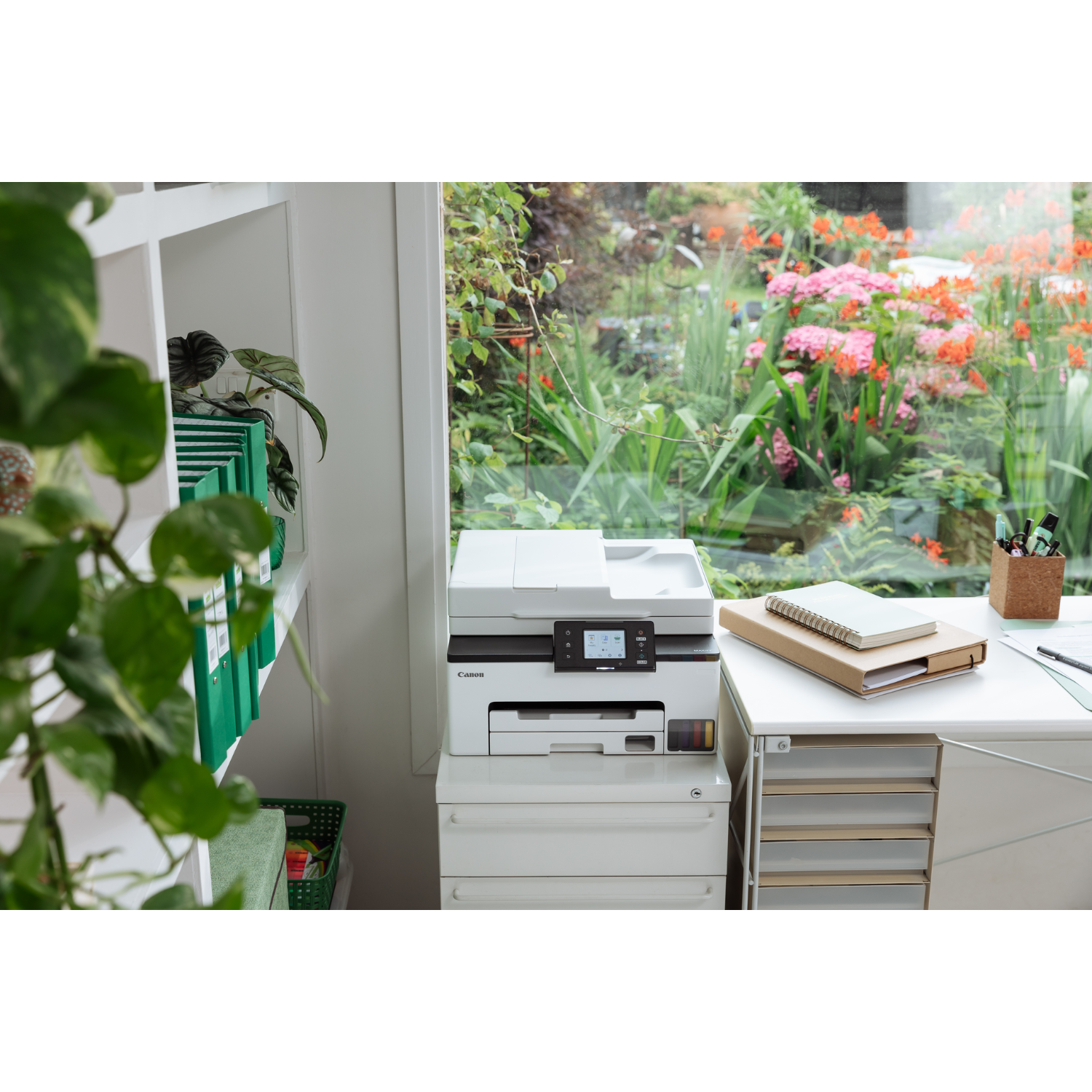 Drukarka na białym biurku obok okna. Widoczne również papiery, notatnik i rośliny.