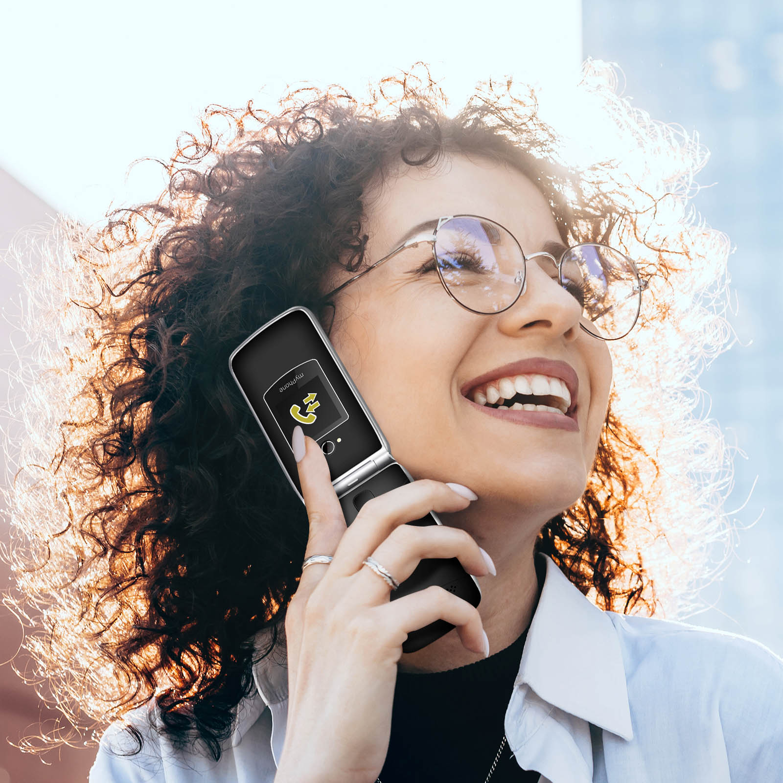 Mujer con gafas sonriendo mientras sostiene un teléfono plegable en su oído con el sol de fondo.