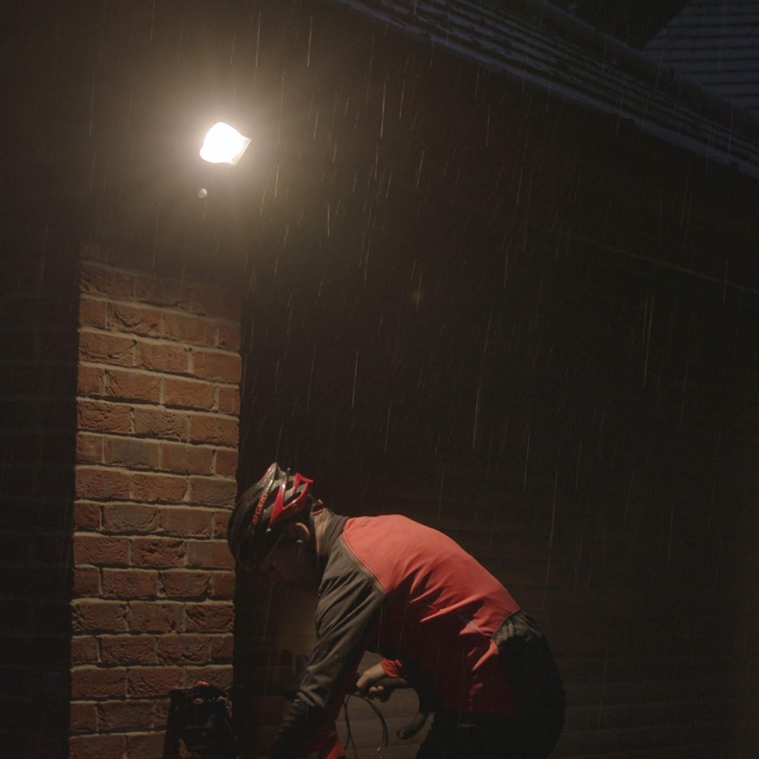 Hombre montando en bicicleta bajo la lluvia por la noche, bajo una luz brillante y cuadrada en una pared de ladrillo.