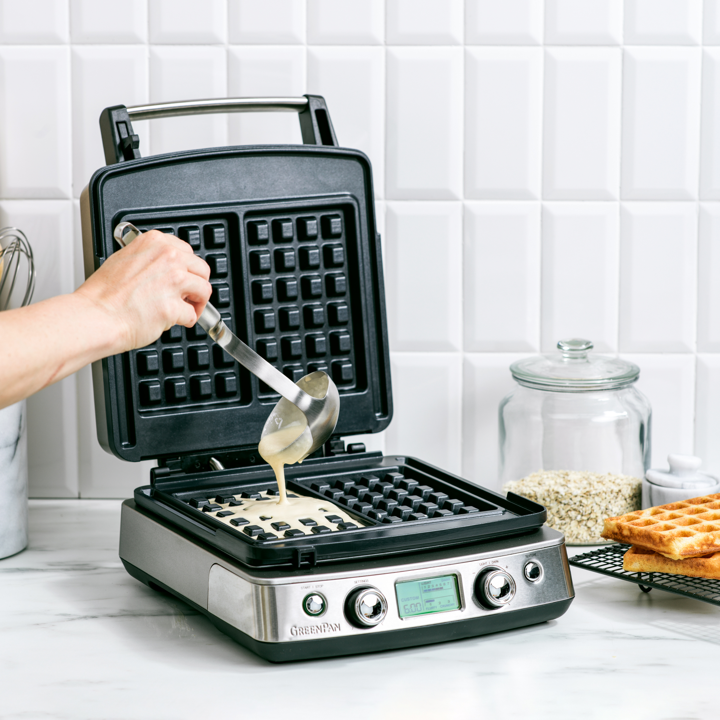 Person gießt Teig in einen Waffeleisen. Waffeln auf einem Rost. Weiße Küchenumgebung.