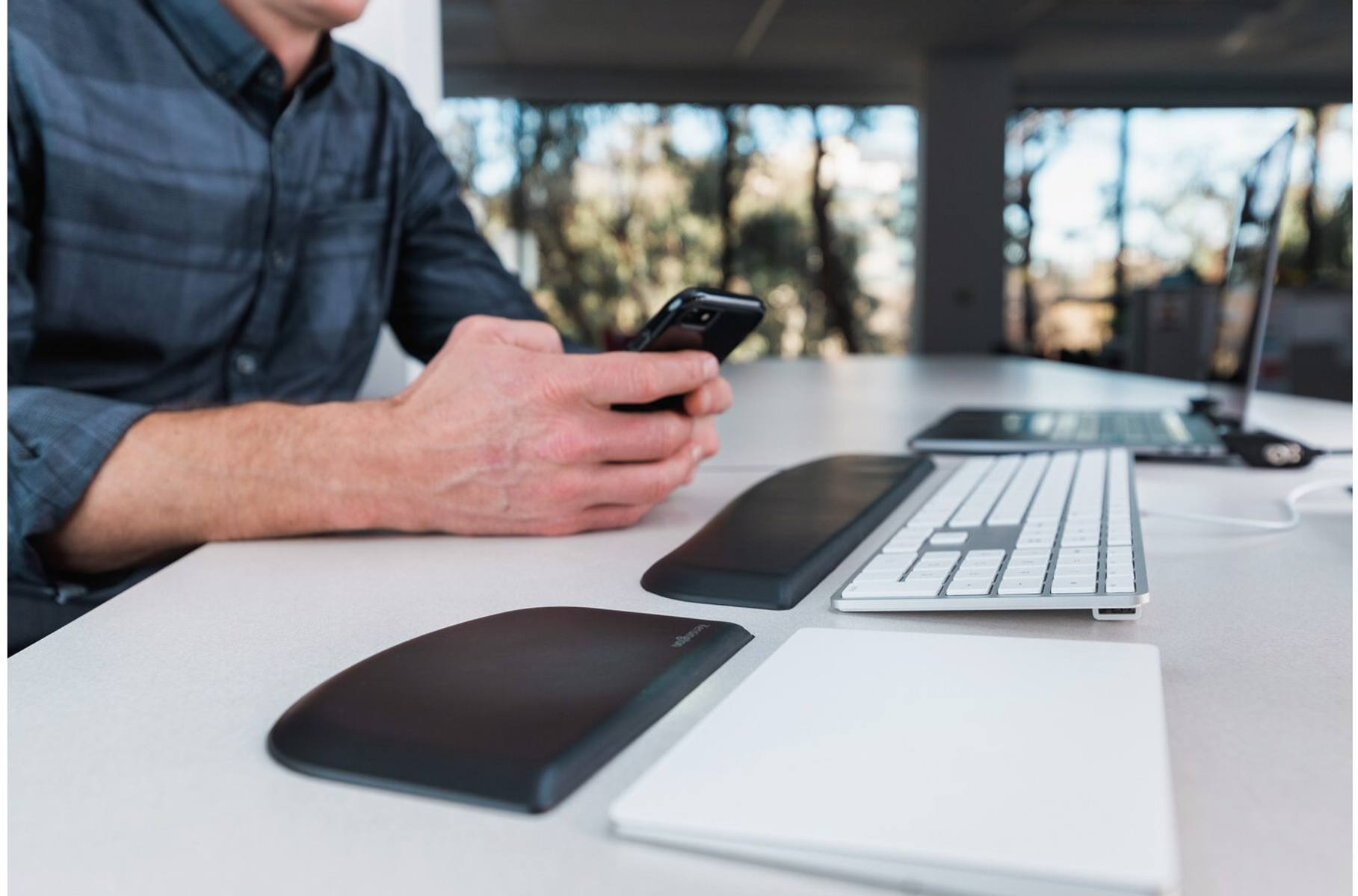 Un homme utilise un smartphone, avec un clavier et des accessoires sur un bureau. Arrière-plan : fenêtre et arbres.