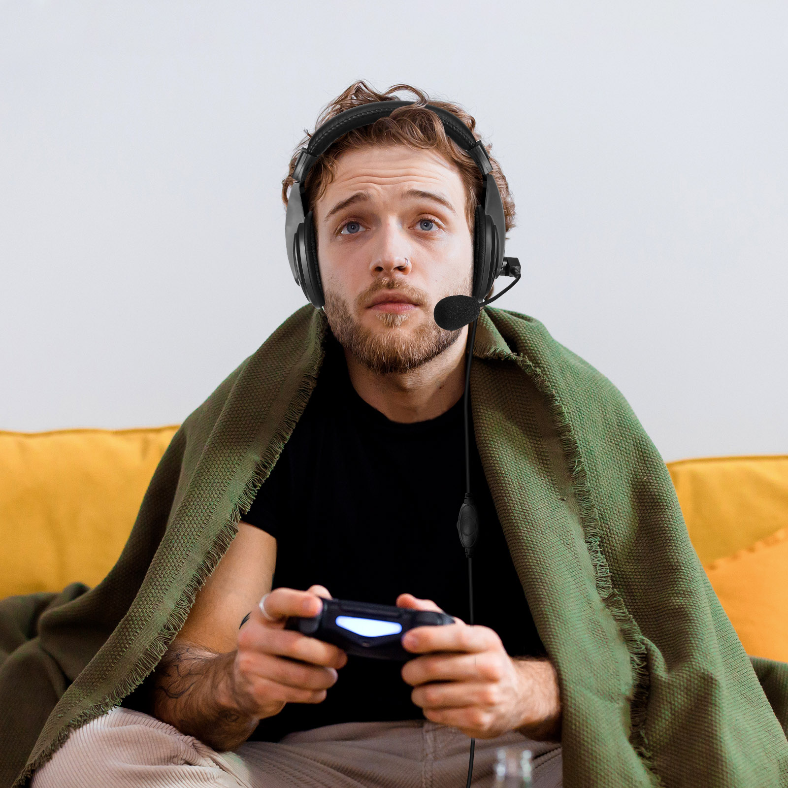 Jeune homme portant un casque, tenant une manette de jeu, couvert d'une couverture verte, dans une pièce jaune.