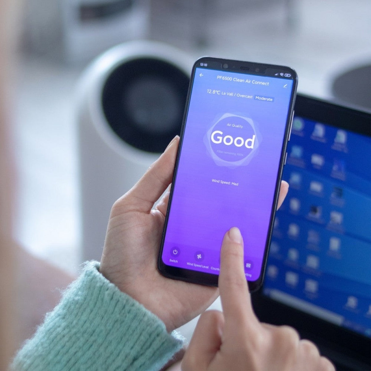 Una mano che tiene uno smartphone che mostra "Buono" per la qualità dell'aria. Un laptop è sullo sfondo.