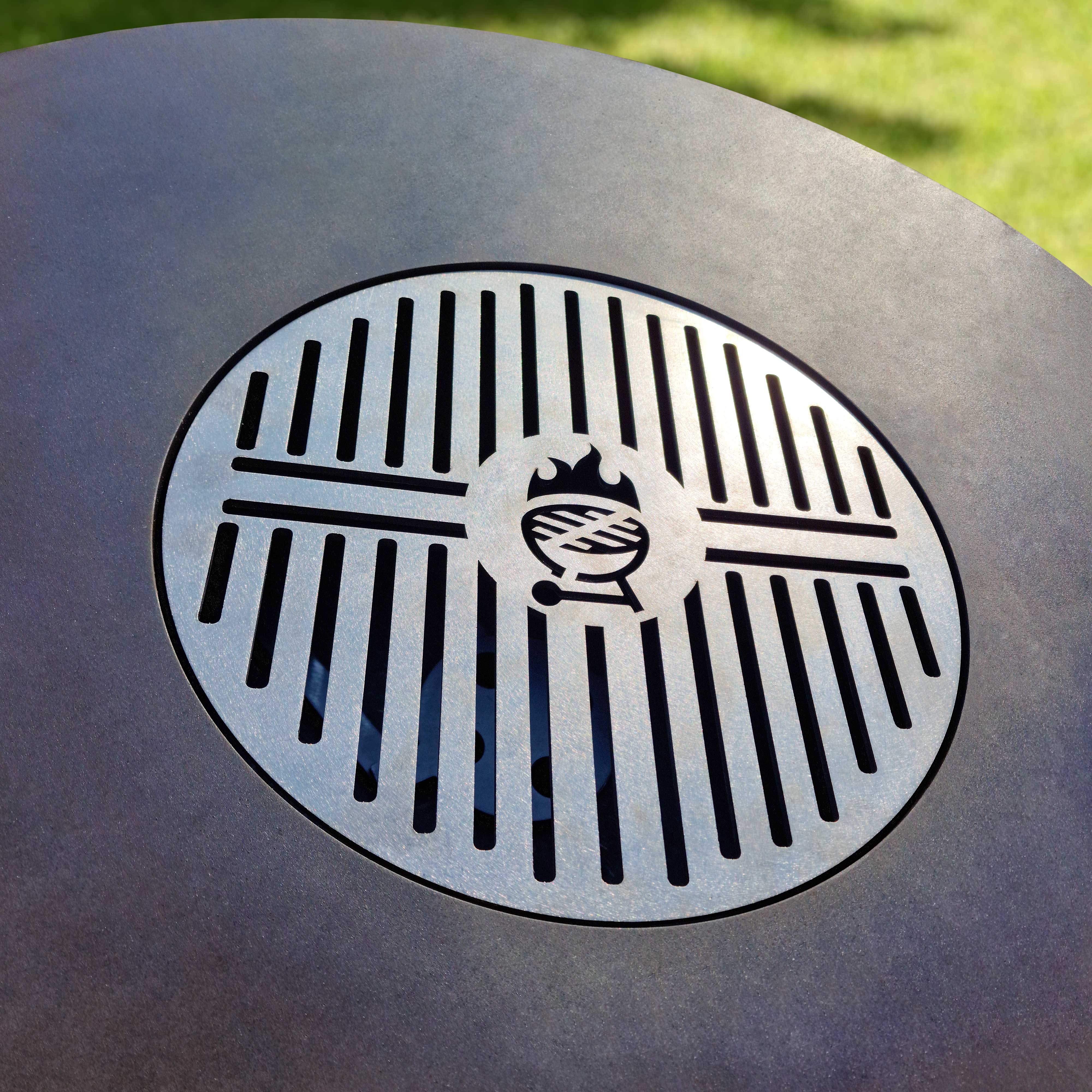 Metallgrill mit Grillsymbol in der Mitte, auf dunkler Oberfläche, mit grünem Gras im Hintergrund.