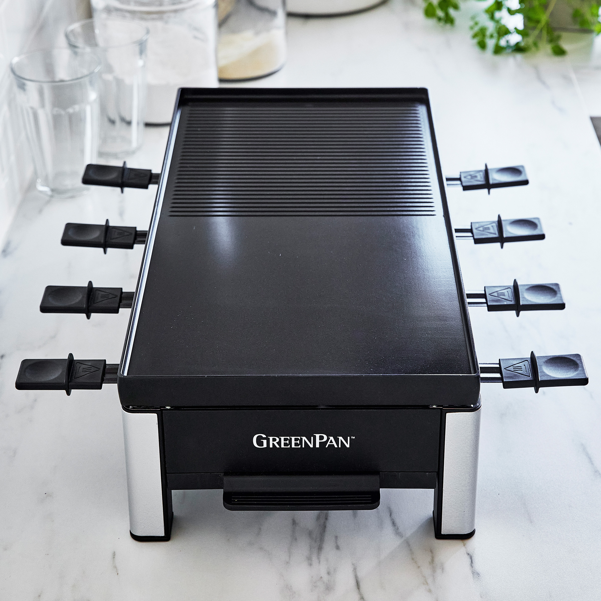Schwarzer Raclette-Grill mit Griffen und silbernen Beinen, mit Gläsern und Gläsern im Hintergrund.
