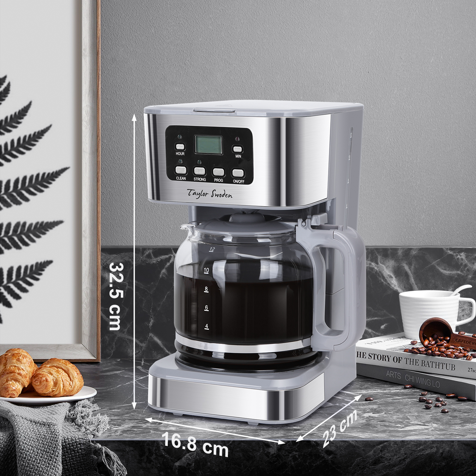 Macchina da caffè argento e grigia su un piano di lavoro con croissant e un libro, con dimensioni.