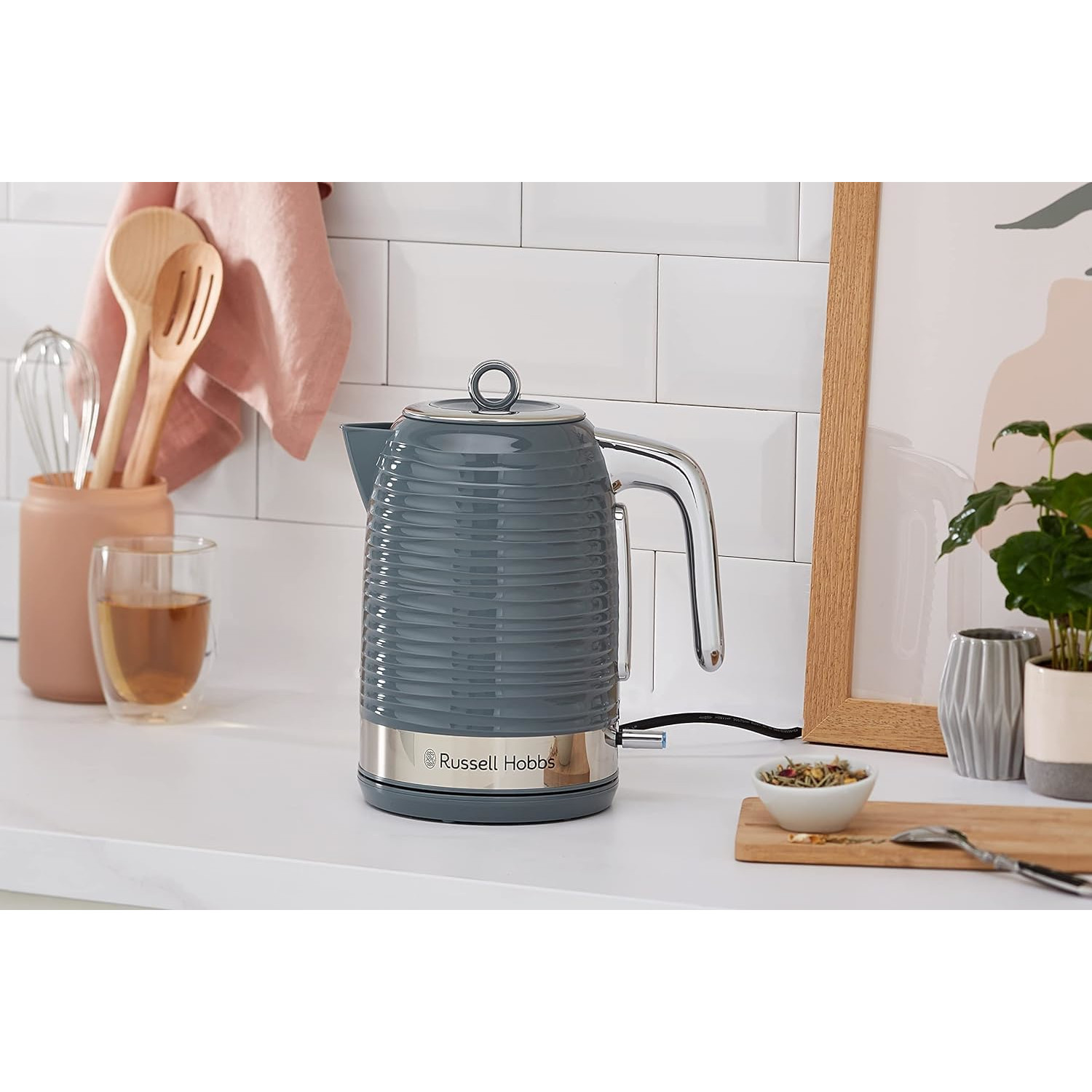 Un bollitore grigio Russell Hobbs su un piano di lavoro bianco accanto a utensili di legno e una pianta.