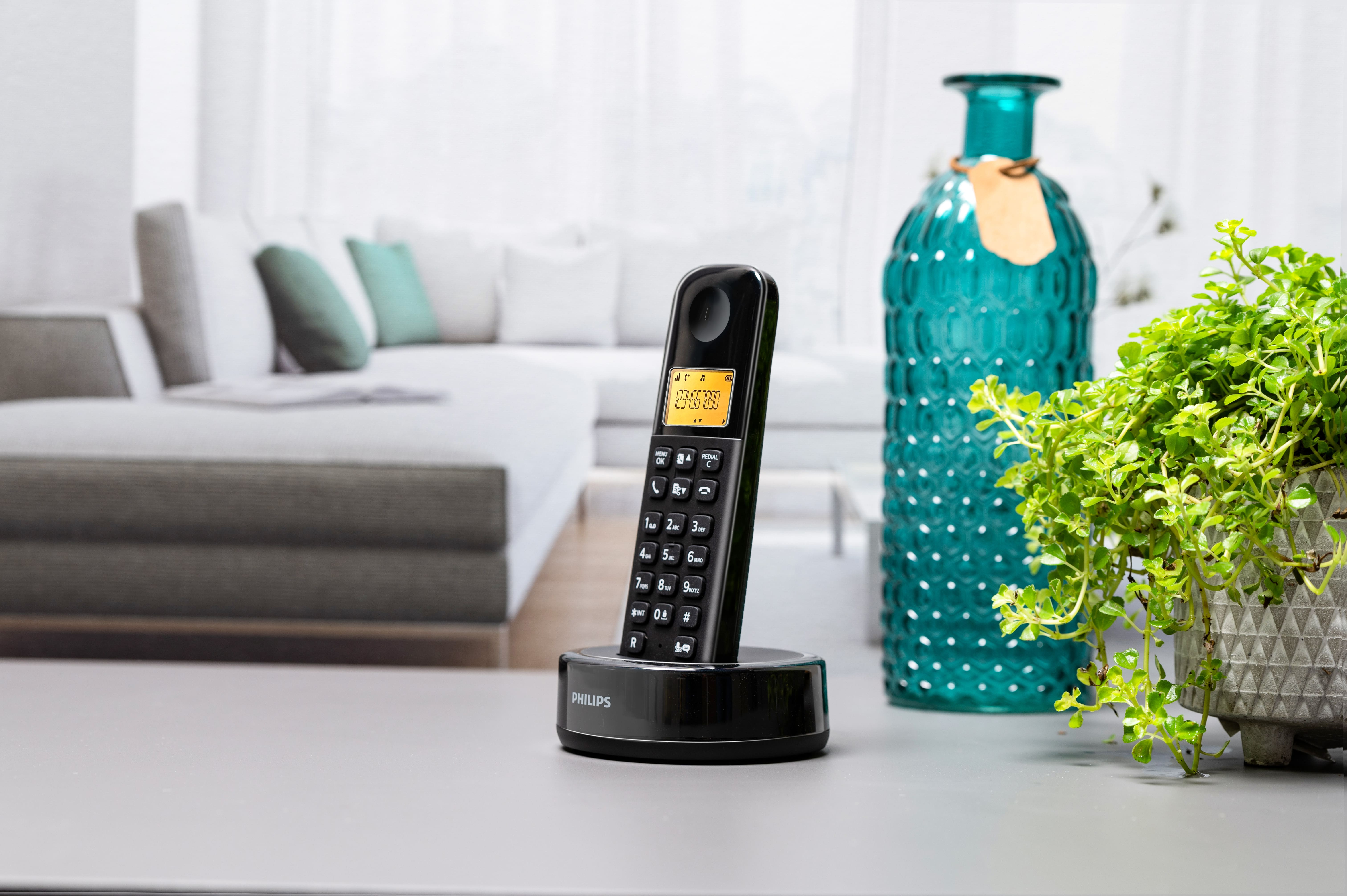 Téléphone sans fil noir sur une base de chargement, une bouteille verte et une plante verte. Fond clair.