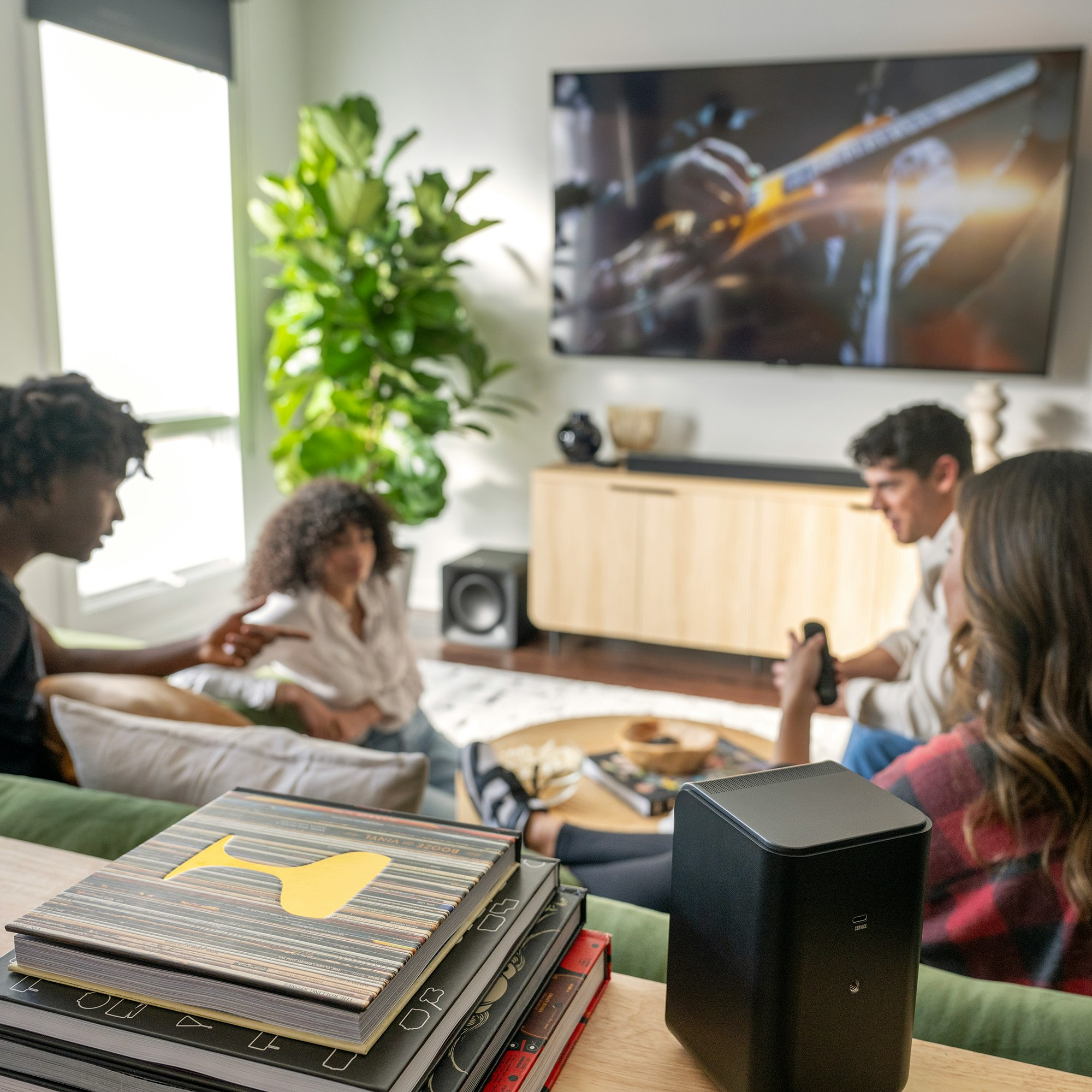Vier Personen sitzen und unterhalten sich in einem Wohnzimmer. Bücher, Fernseher und Lautsprecher.