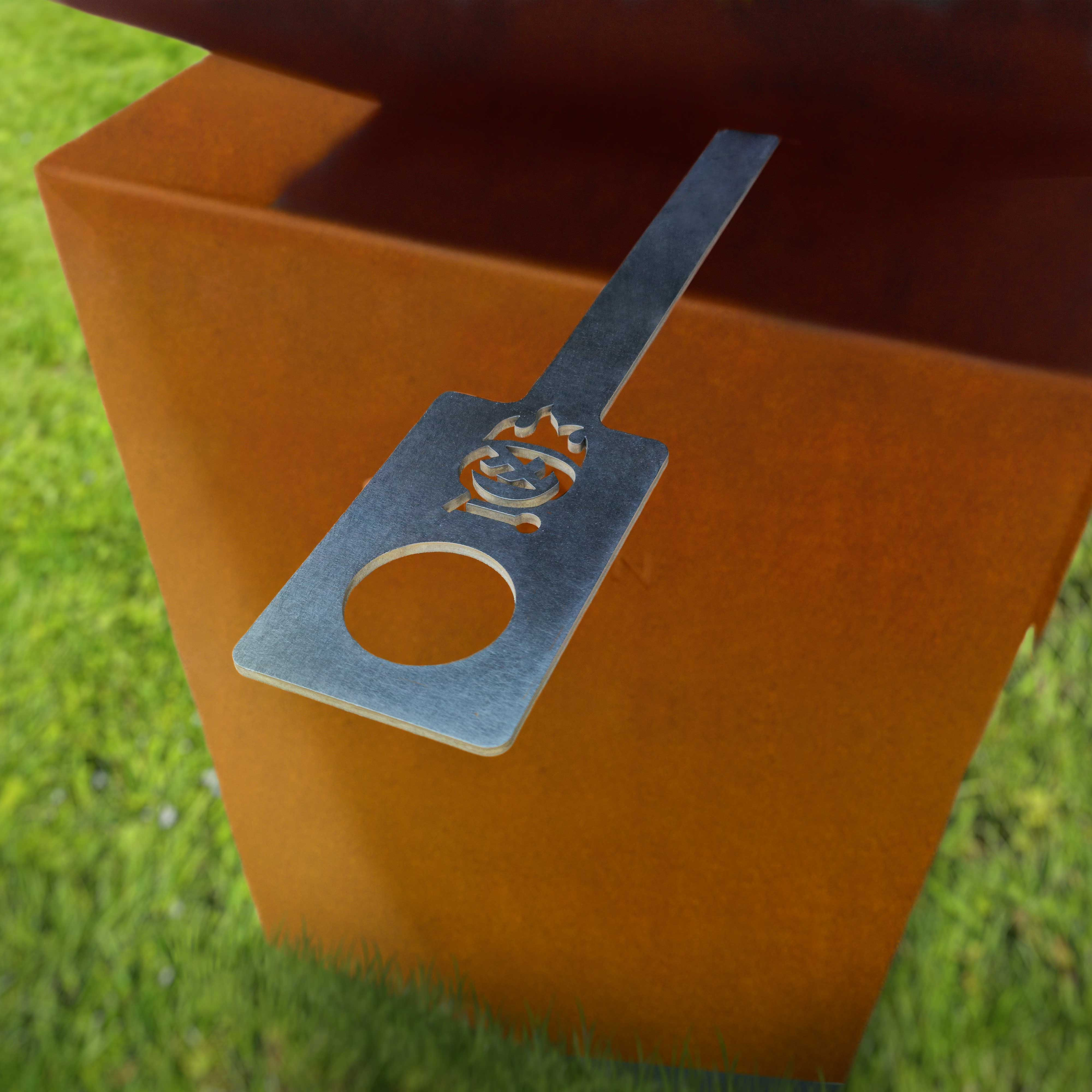 Ein Metallspatel mit Feuerlogo, der auf einer orangefarbenen Metallbox auf Gras liegt.