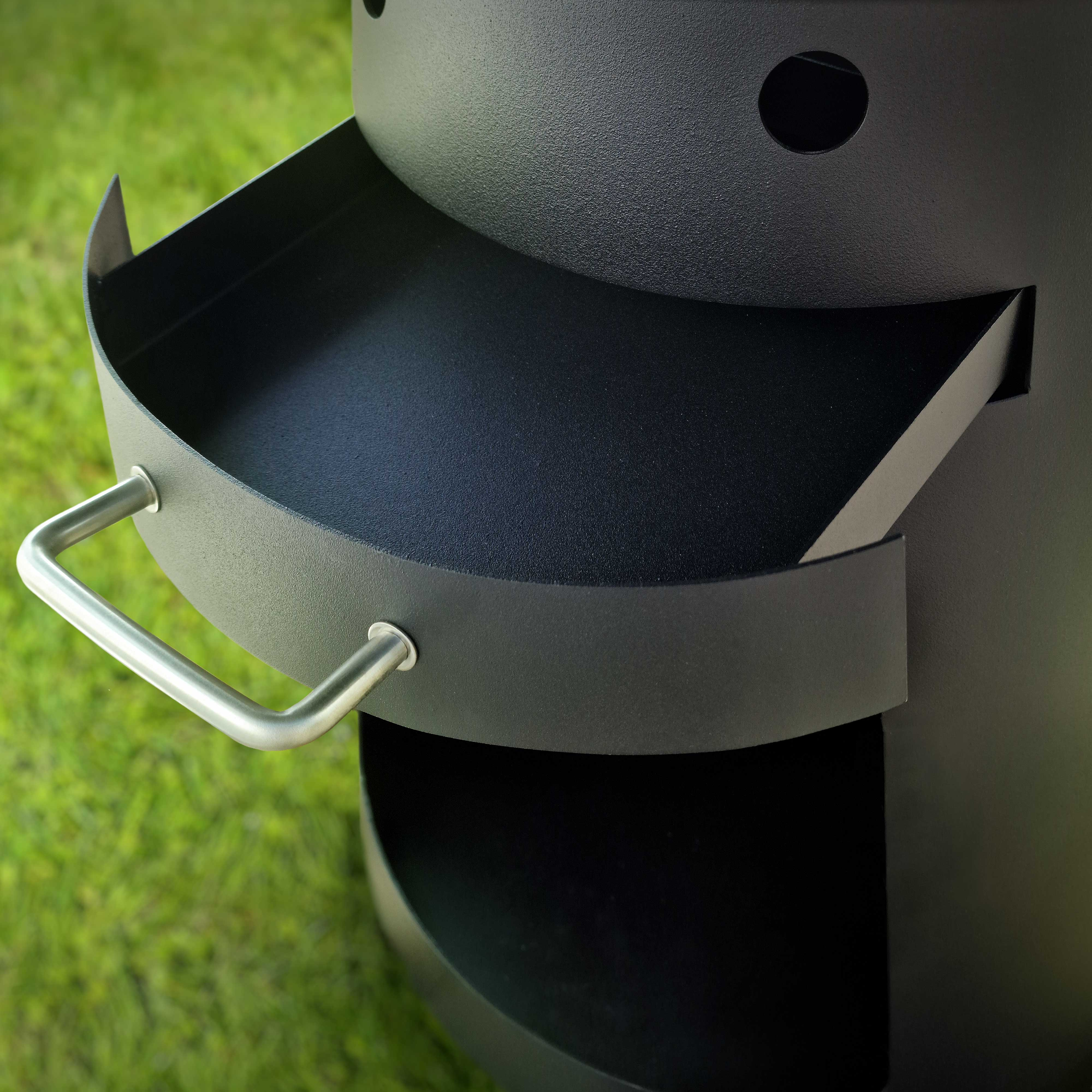 Ein metallischer Aschenbecher eines Grills wird gezeigt. Er ist schwarz und hat einen Griff.